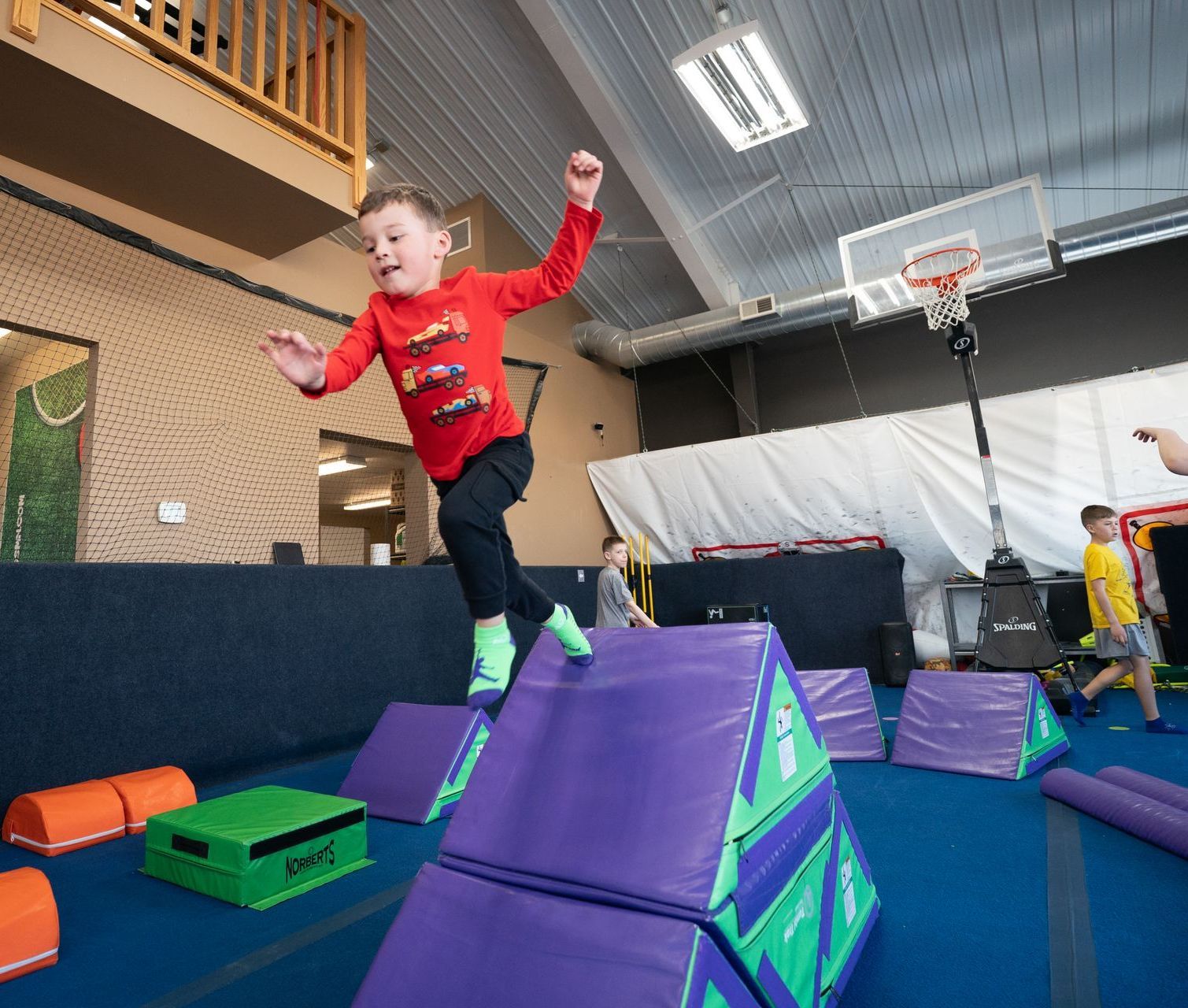 Child jumping on foam obstacles in an indoor play gym with a trampoline and climbing structures.