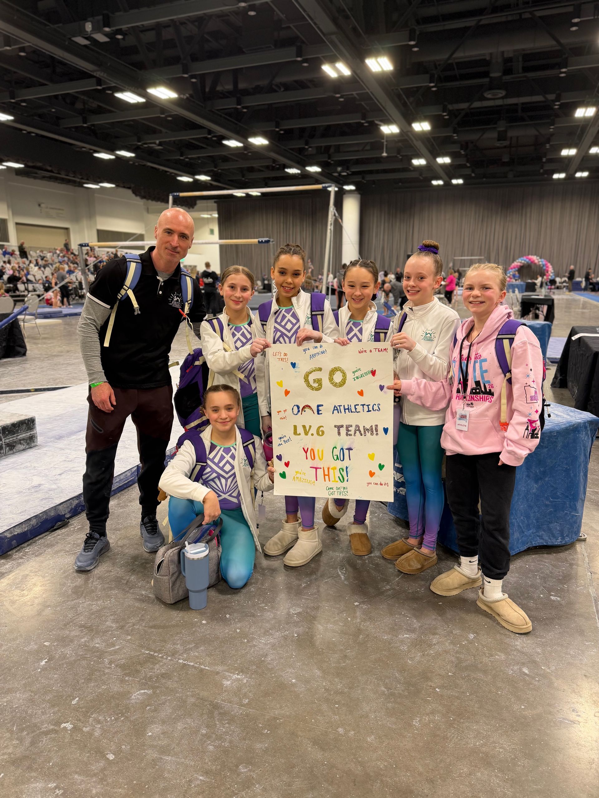 Group of young athletes and coach holding a colorful sign at an indoor sports venue.