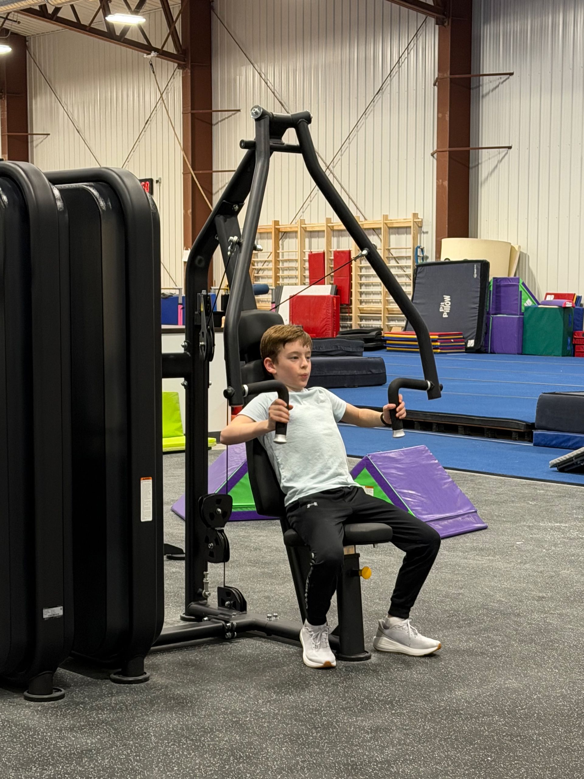 Child exercising on a seated machine in a gym, with black equipment and purple mats nearby.