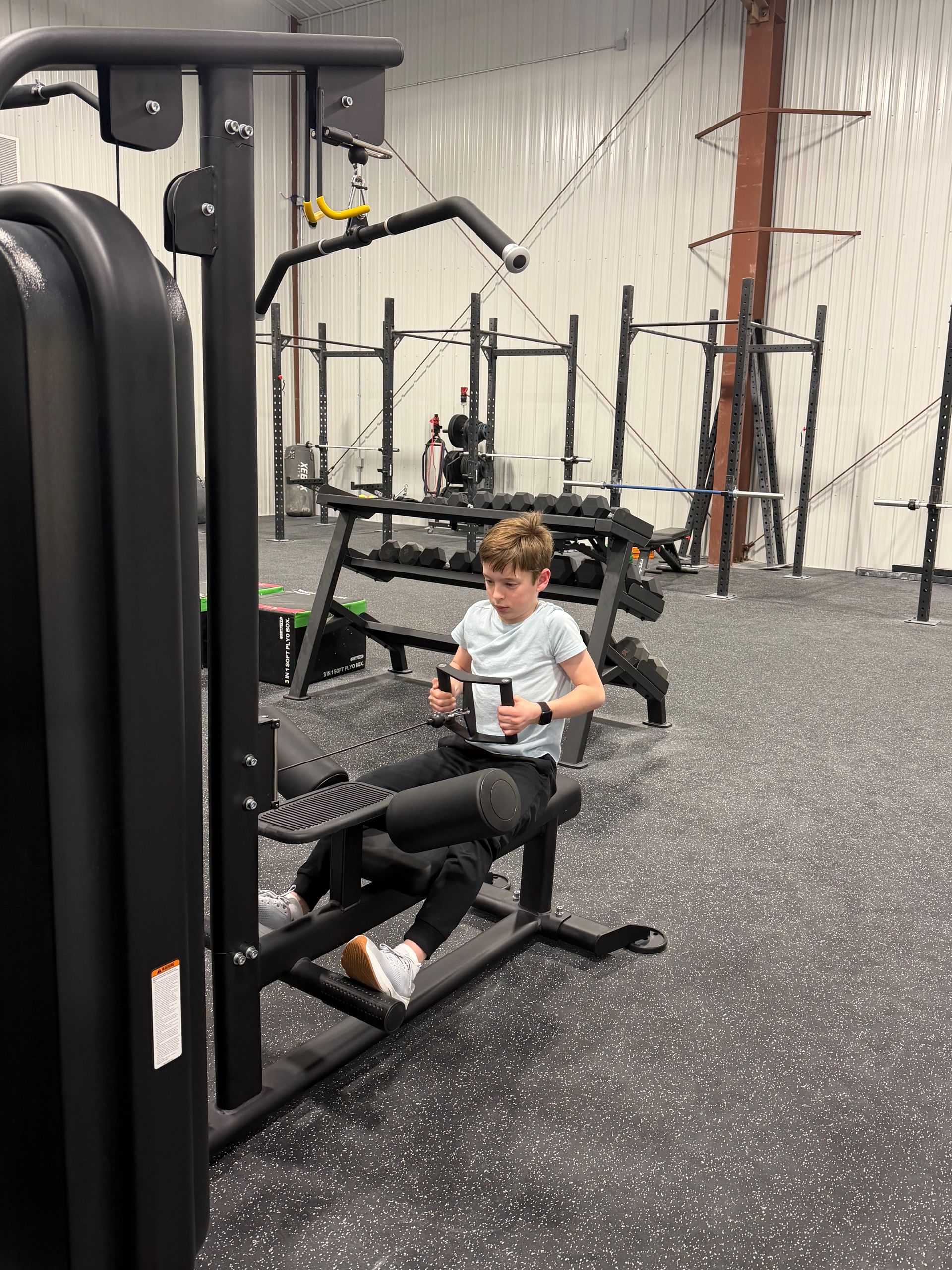 Child seated on a black gym machine in a weight room with speckled flooring and exercise equipment in the background