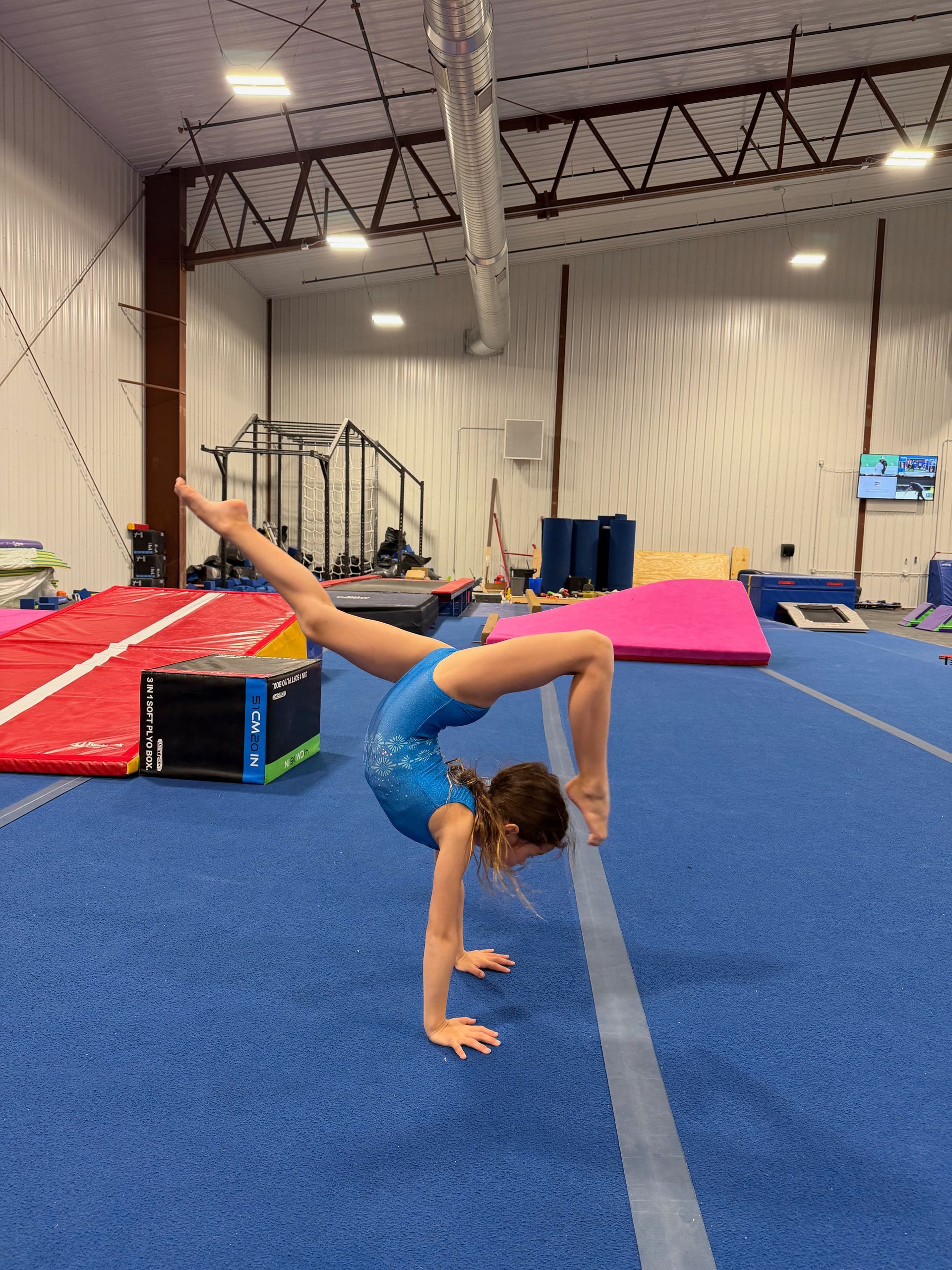 Gymnast balancing on one hand in a gym, leg extended upward on blue mats.
