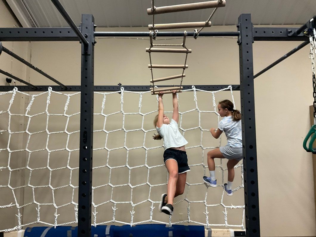 Two climbers hang from a rope ladder obstacle in an indoor gym.