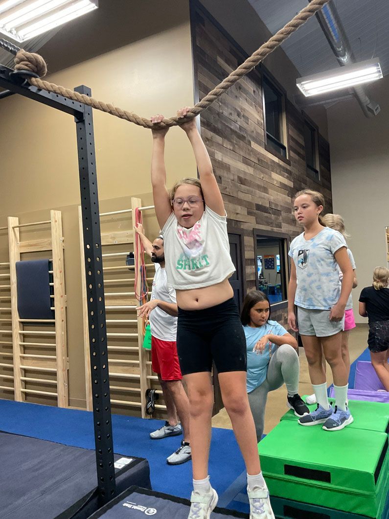 Child hanging from rope in an indoor gym while others stand nearby on padded platforms