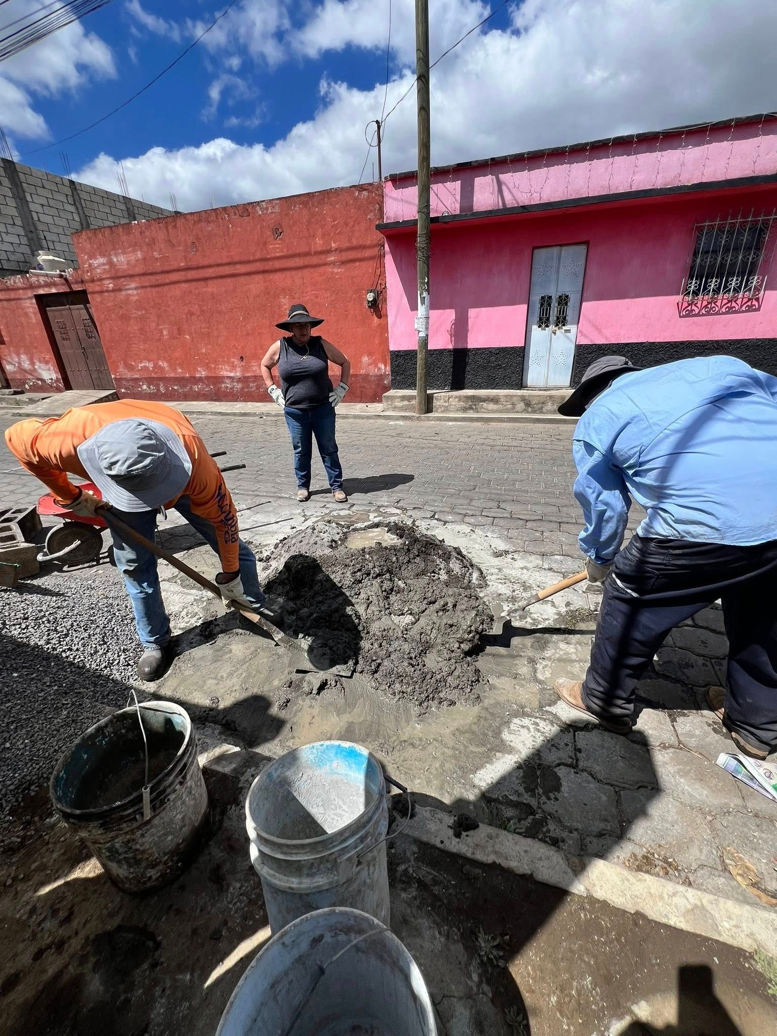 Three people work on a street repair, mixing cement in a hole between a red and a pink building under a sunny sky.