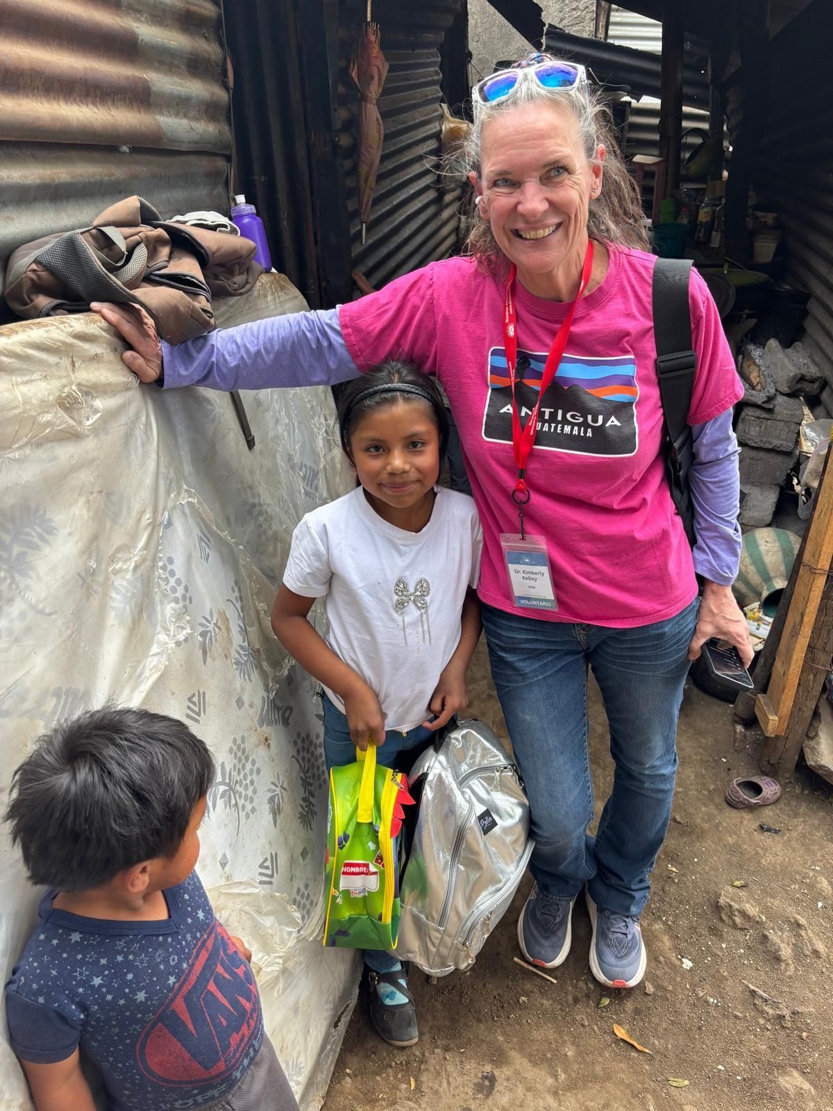 A smiling volunteer in a pink shirt and blue jeans stands with two children holding backpacks in a simple outdoor setting.