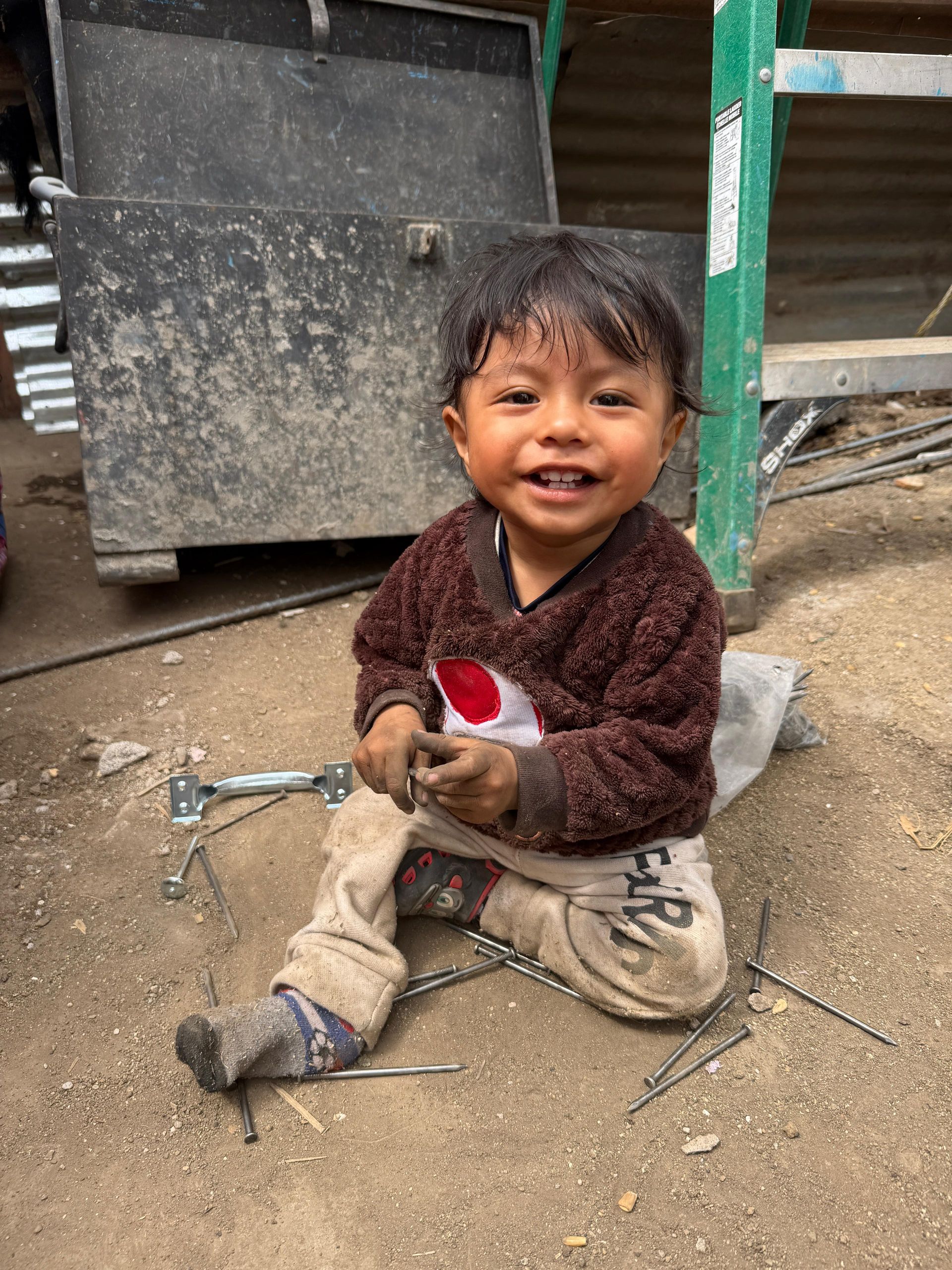 A young child with a happy expression sits on a dirt floor, wearing a brown fuzzy sweater and light pants.