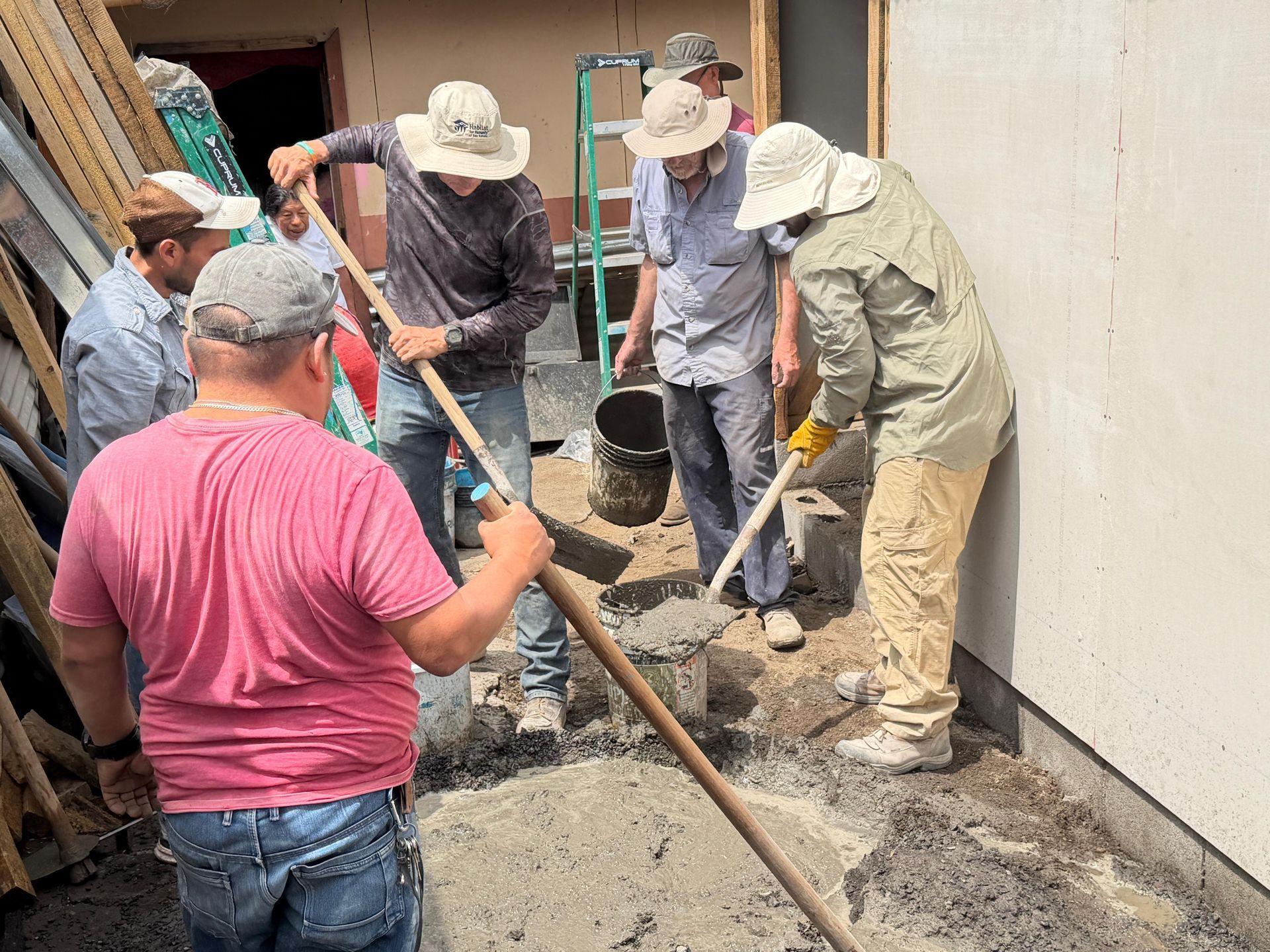 Six people wearing hats work together to mix and spread wet cement on the ground at a construction site.