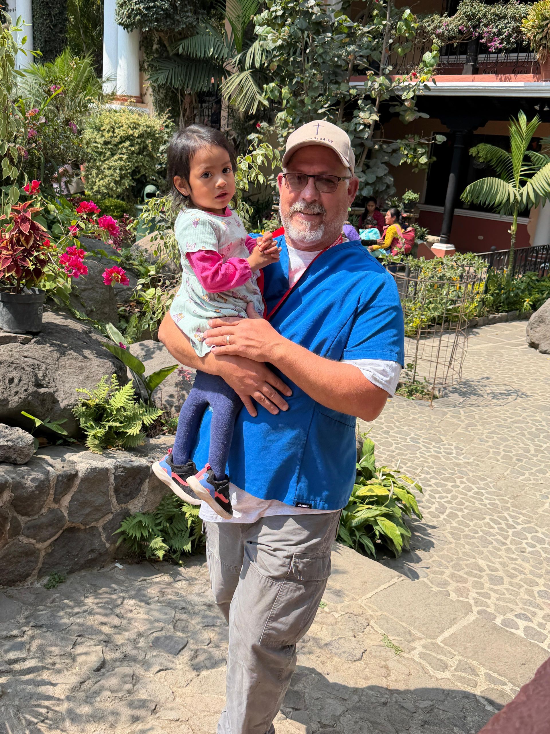 A person in a blue scrub top and cap holds a young child in their arms, standing in a sunny, landscaped garden setting.