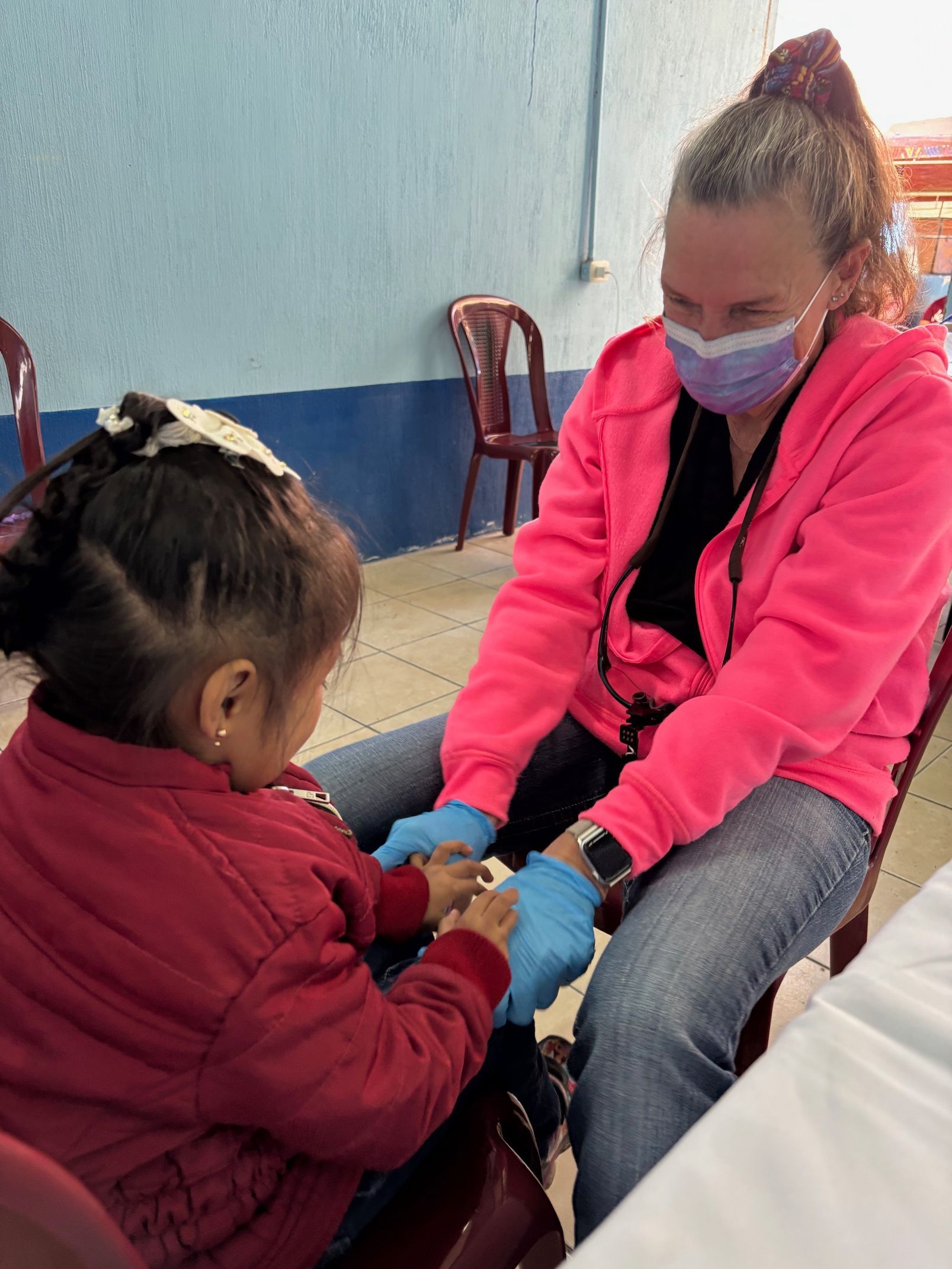 A healthcare worker in a pink hoodie and mask holds a young patient's hands while providing care in a clinic setting.