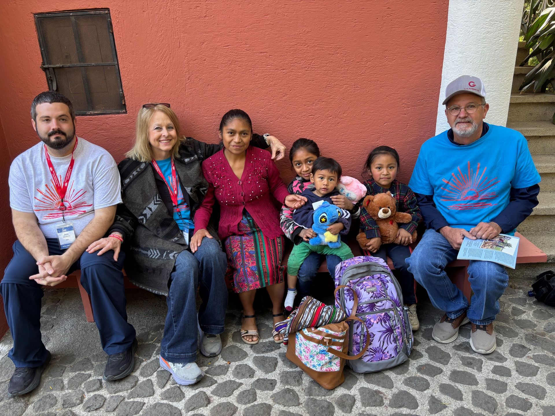 A group of seven people sitting on a bench against a red wall, with children holding stuffed animals and backpacks.