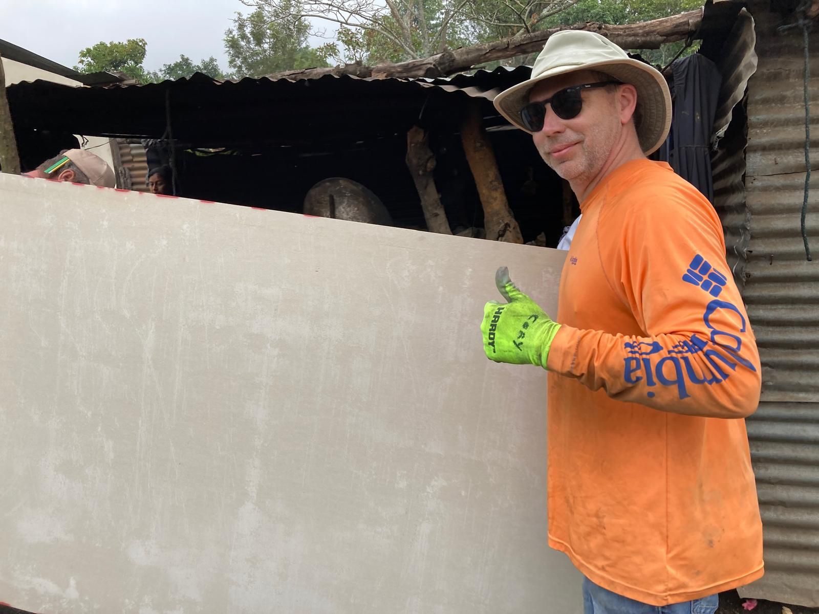 A man wearing an orange shirt and a hat is standing in front of a wall.
