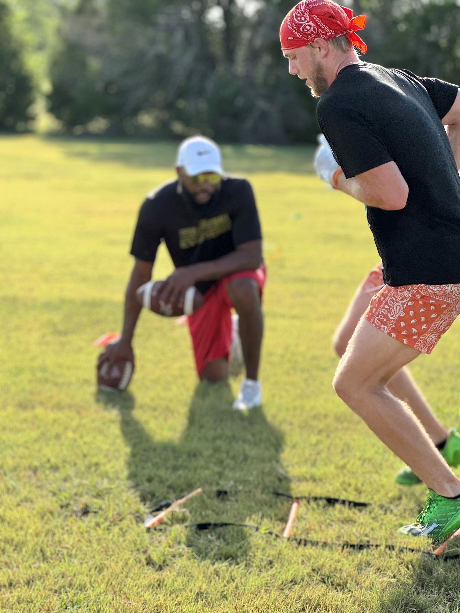 Man in red bandana runs toward kneeling man on grass field during football training.