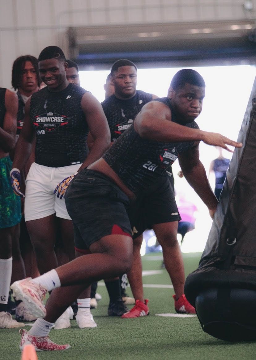 Football player training, pushing a blocking sled. Others watch in a gym, wearing athletic gear.