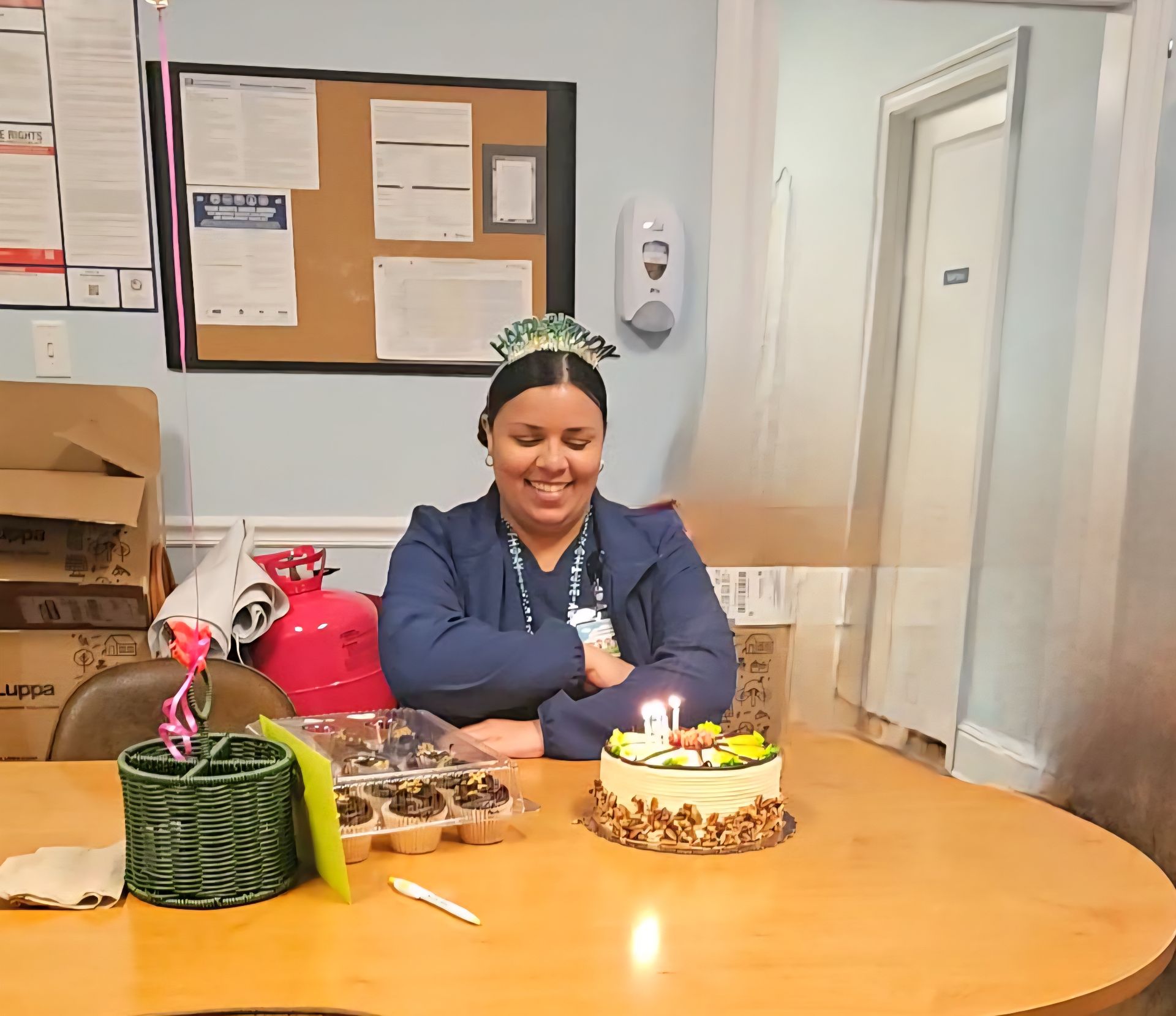 Woman sitting at a table with a birthday cake and cupcakes, smiling. In a room with posters and a door.