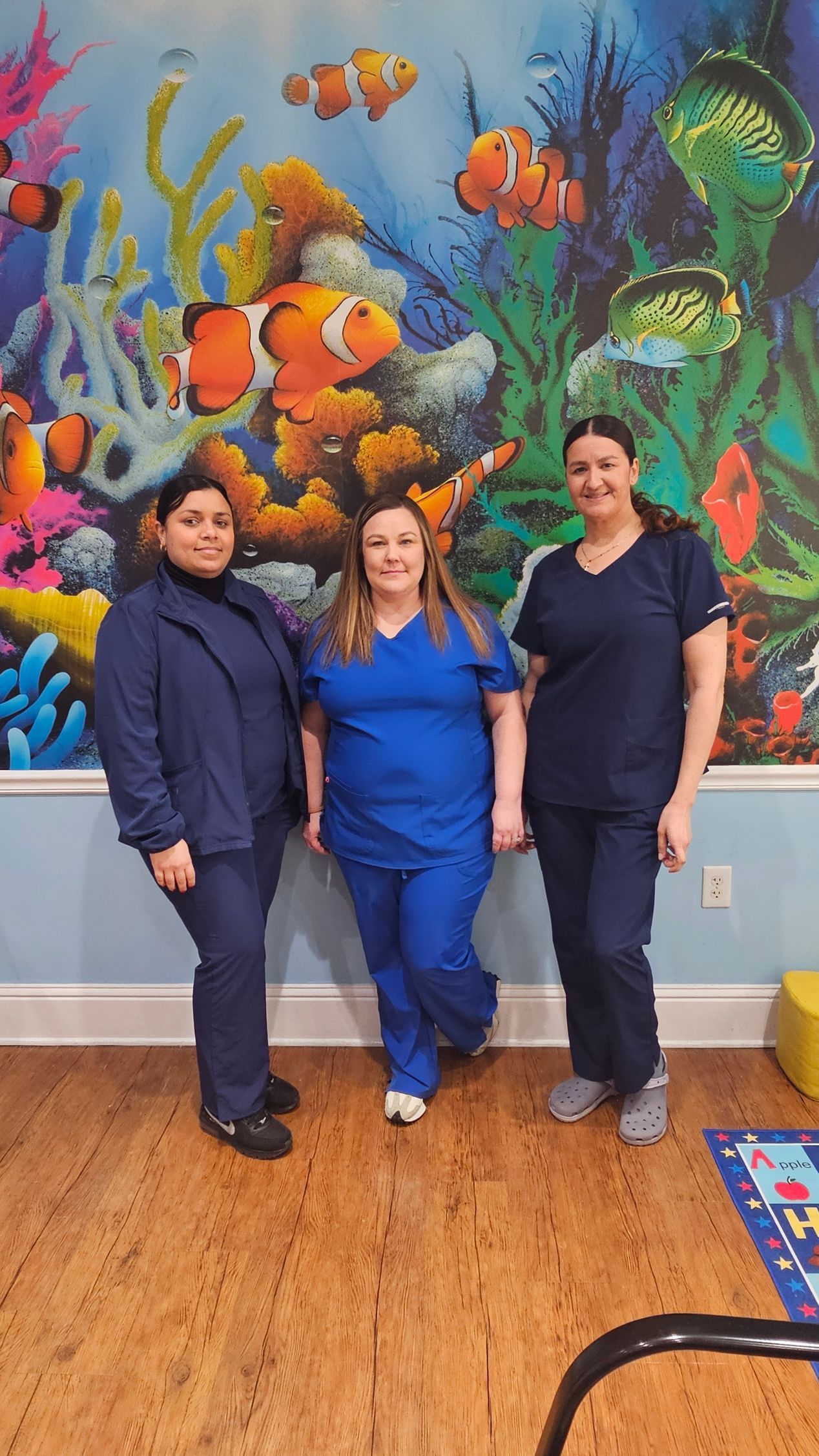 Three people in blue scrubs stand in front of a colorful mural of an underwater scene.