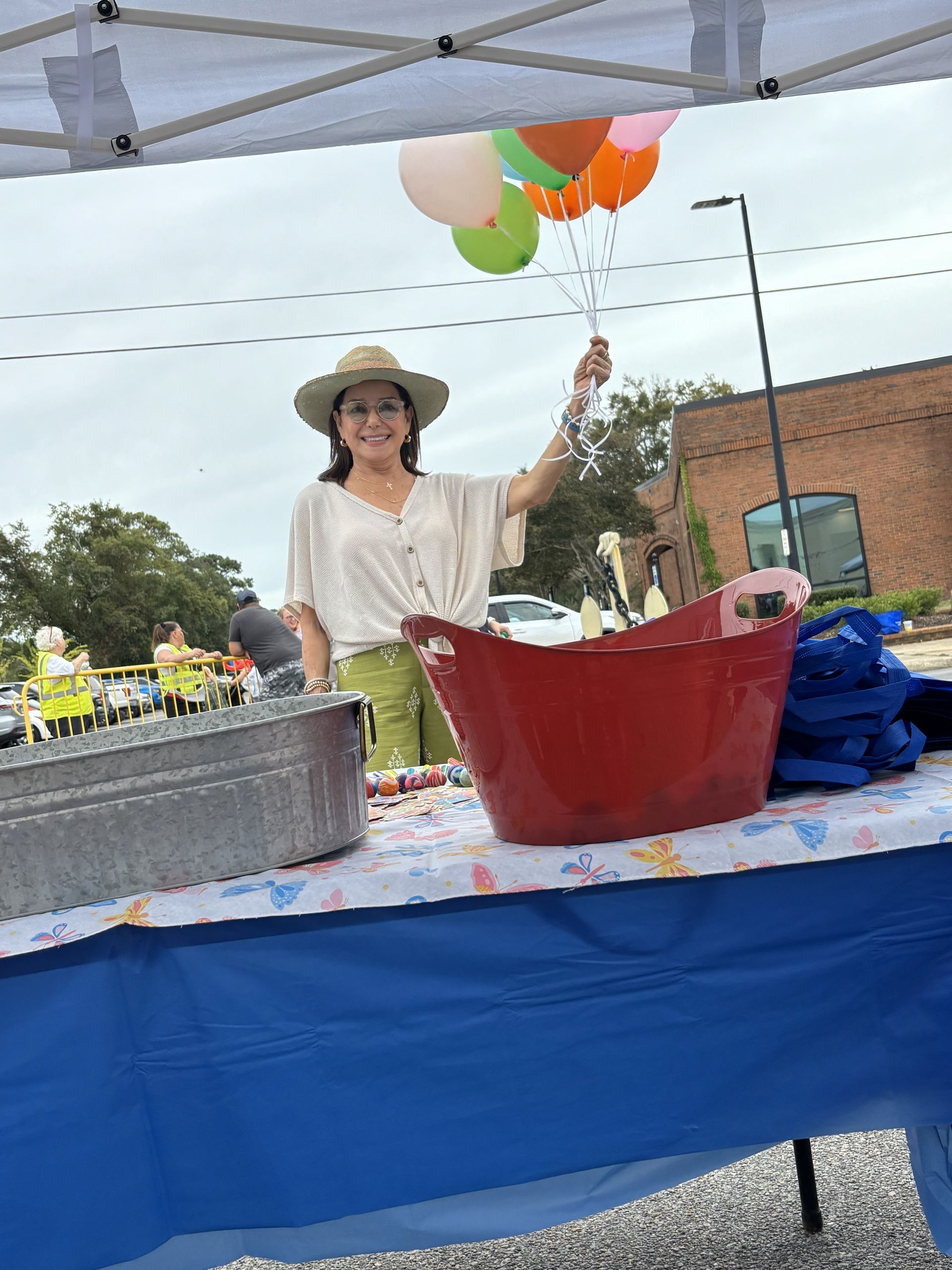Woman under tent holds balloons, smiling at a market table with items, including a red bin.