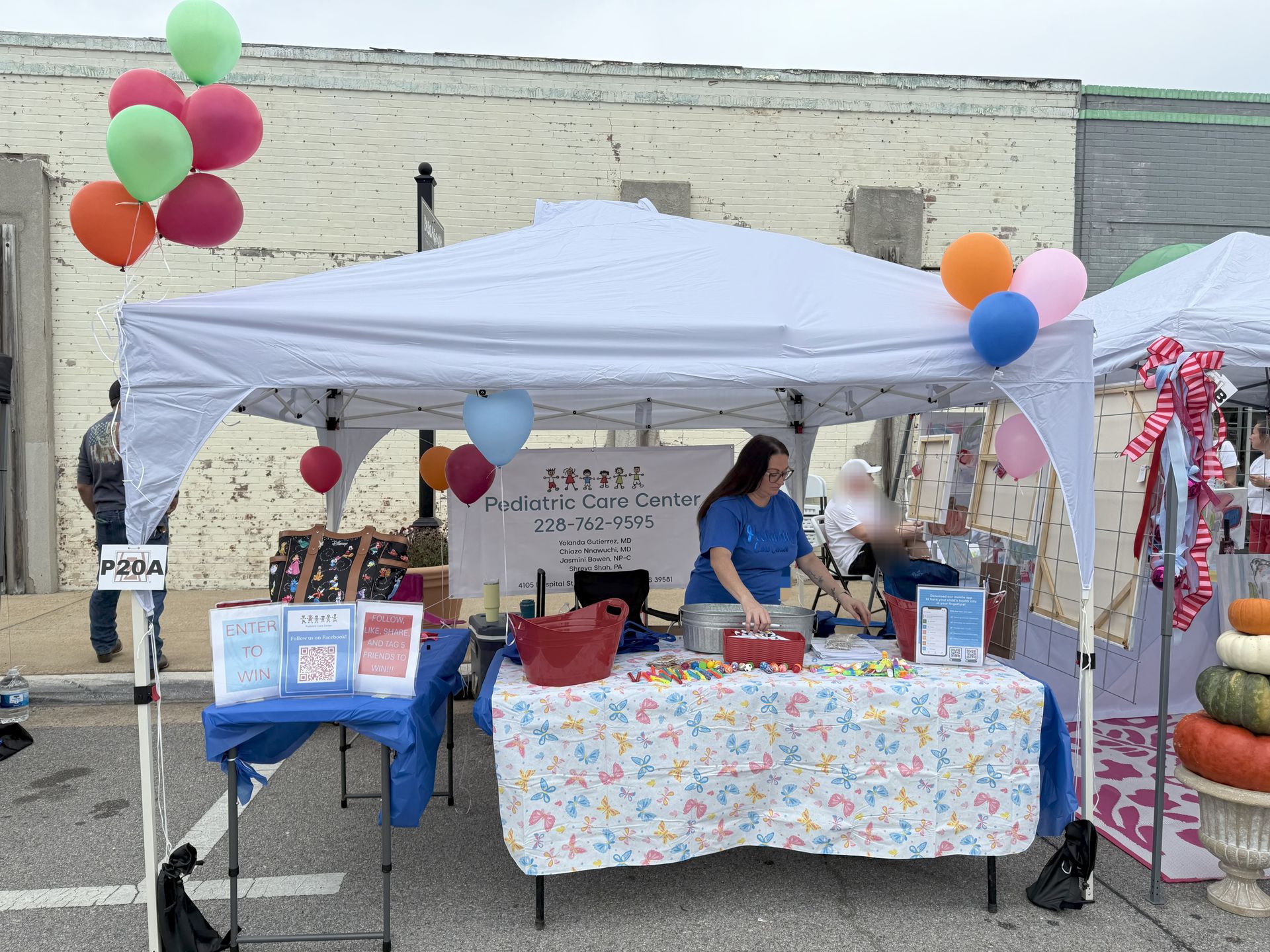 White tent with balloons at a street fair. A person in blue shirt works a table with candy.