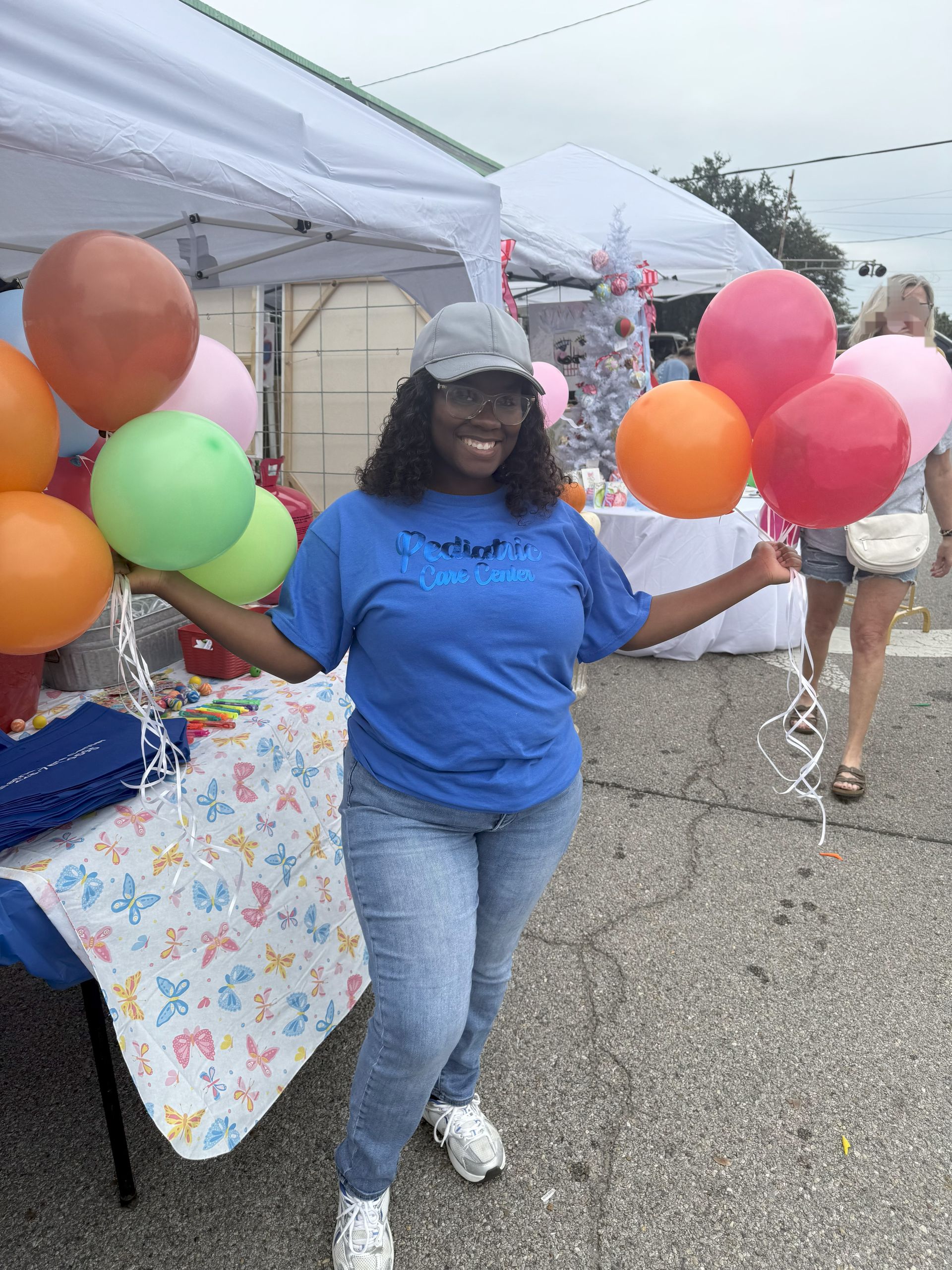 Woman with balloons smiles at outdoor event, wearing blue shirt, jeans, and a cap.