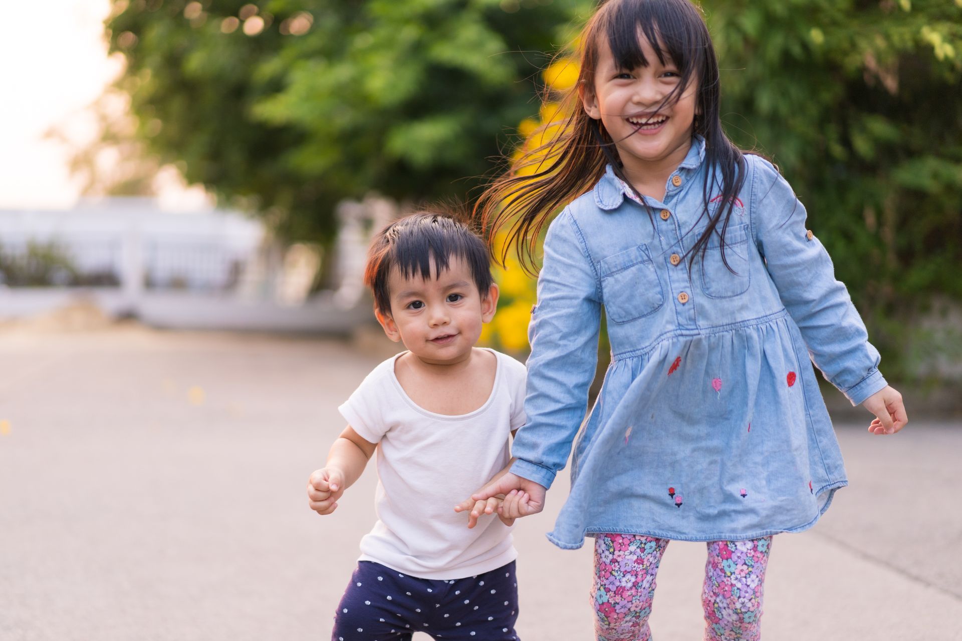 Two children holding hands and smiling outdoors on a sidewalk at sunset.