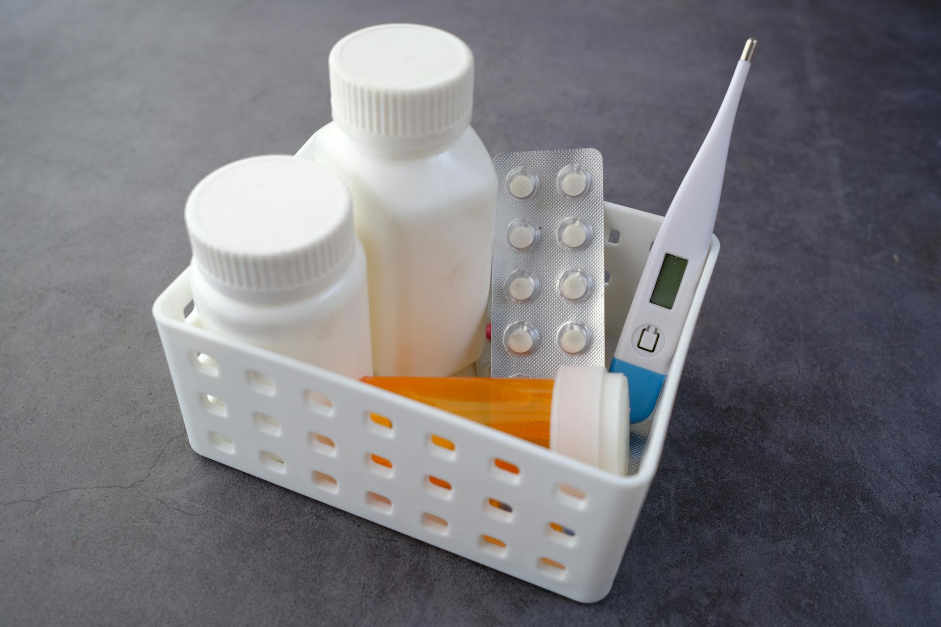 White basket holding medicine bottles, blister pack of pills, and a thermometer on a gray surface.
