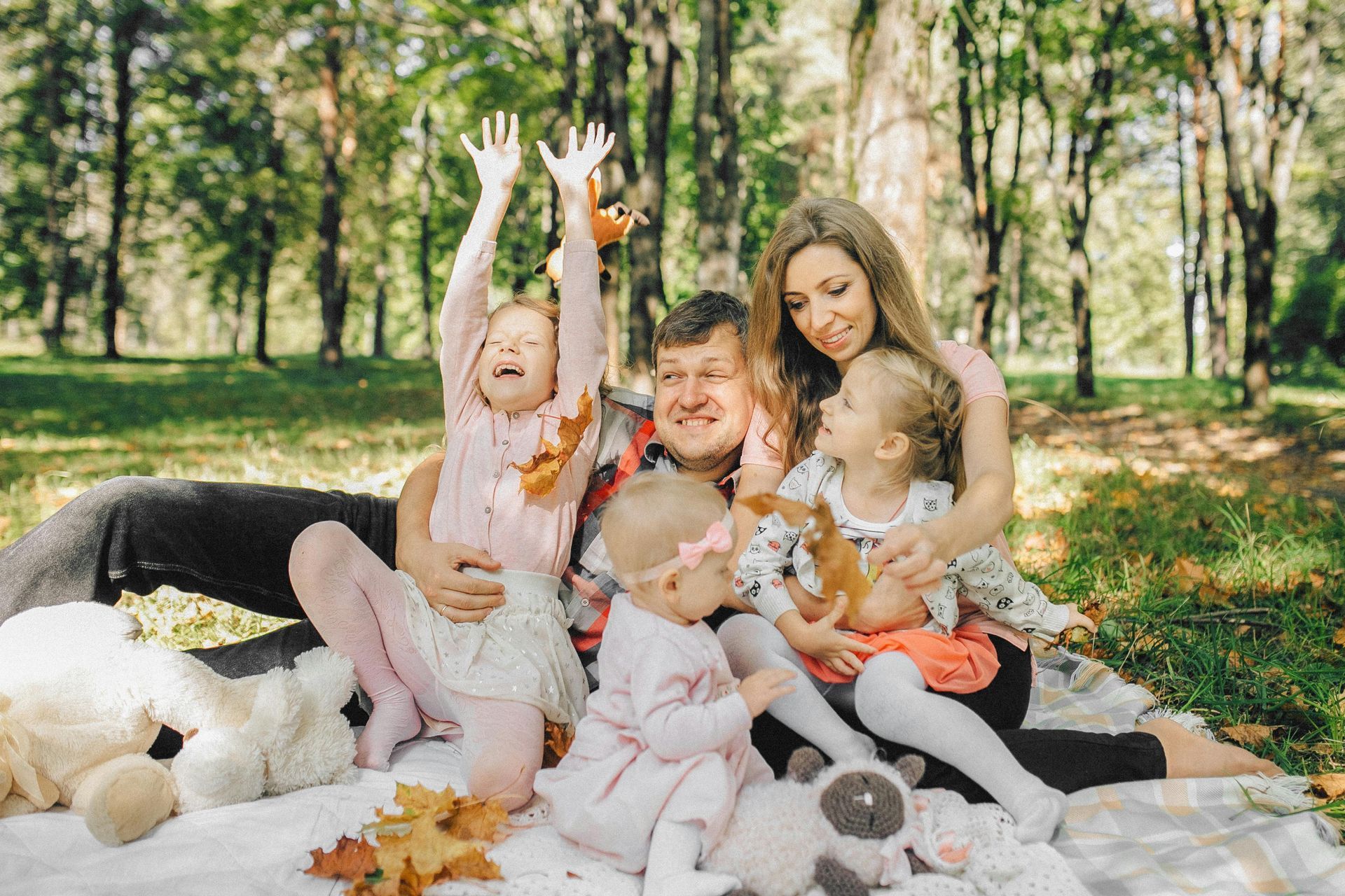 Family sitting on a blanket in a sunlit forest, children holding autumn leaves and smiling.