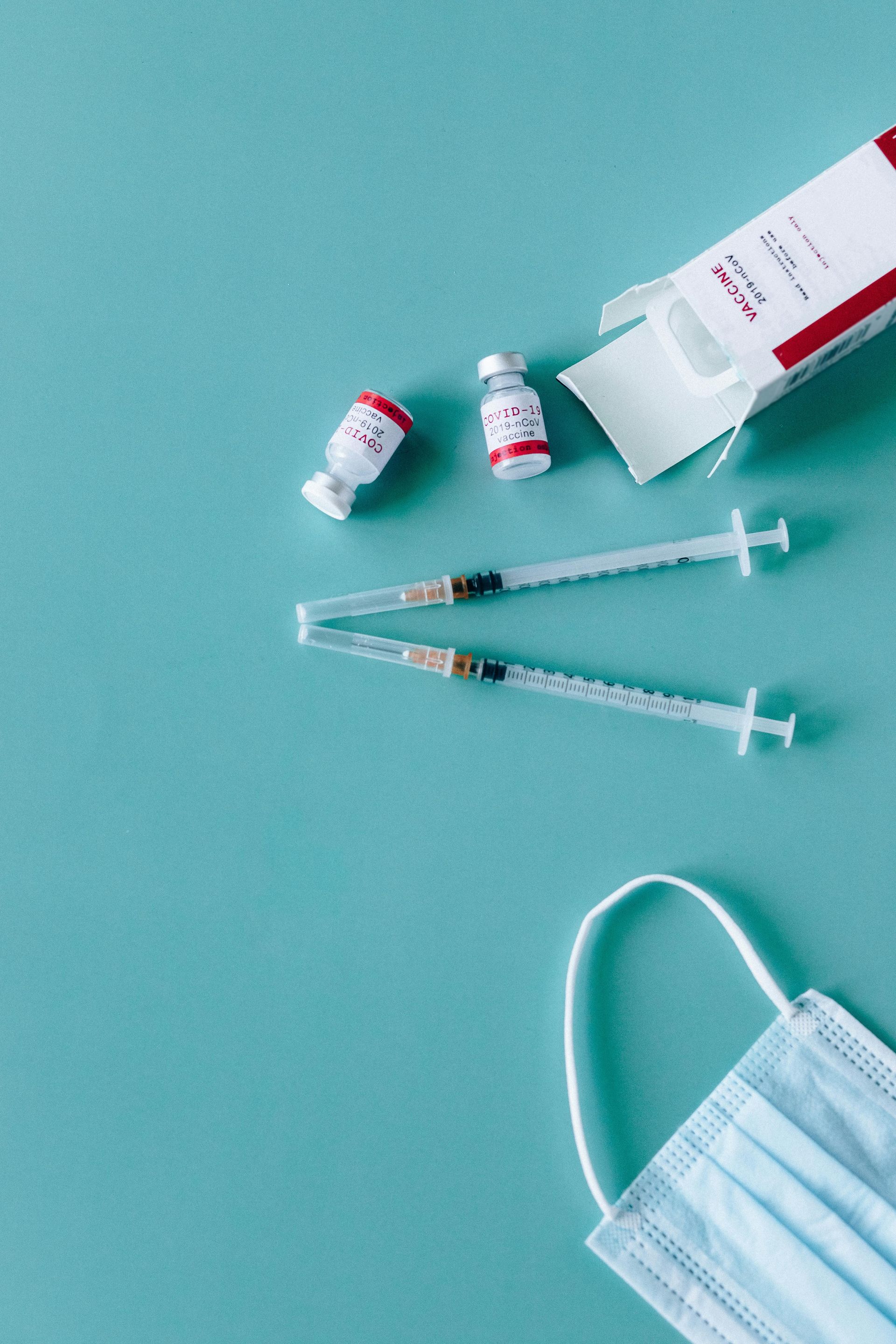 Medical supplies on a teal surface: syringes, vaccine vials, open box, and face mask.