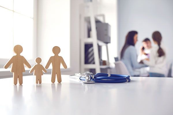 Family figurines beside a stethoscope, with a blurred doctor consulting a child in the background