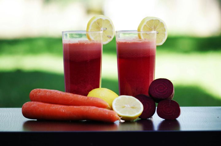 Two glasses of red juice with lemon slices, carrots, lemons, and beets on a table.