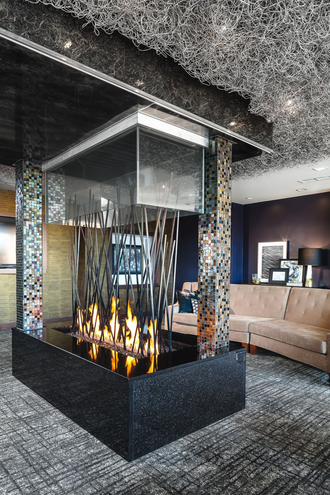 Modern fireplace with black tiled base and glass flames, in a room with matching tile pillars and a beige sofa at The Walkway, which offers Minneapolis apartments for rent.