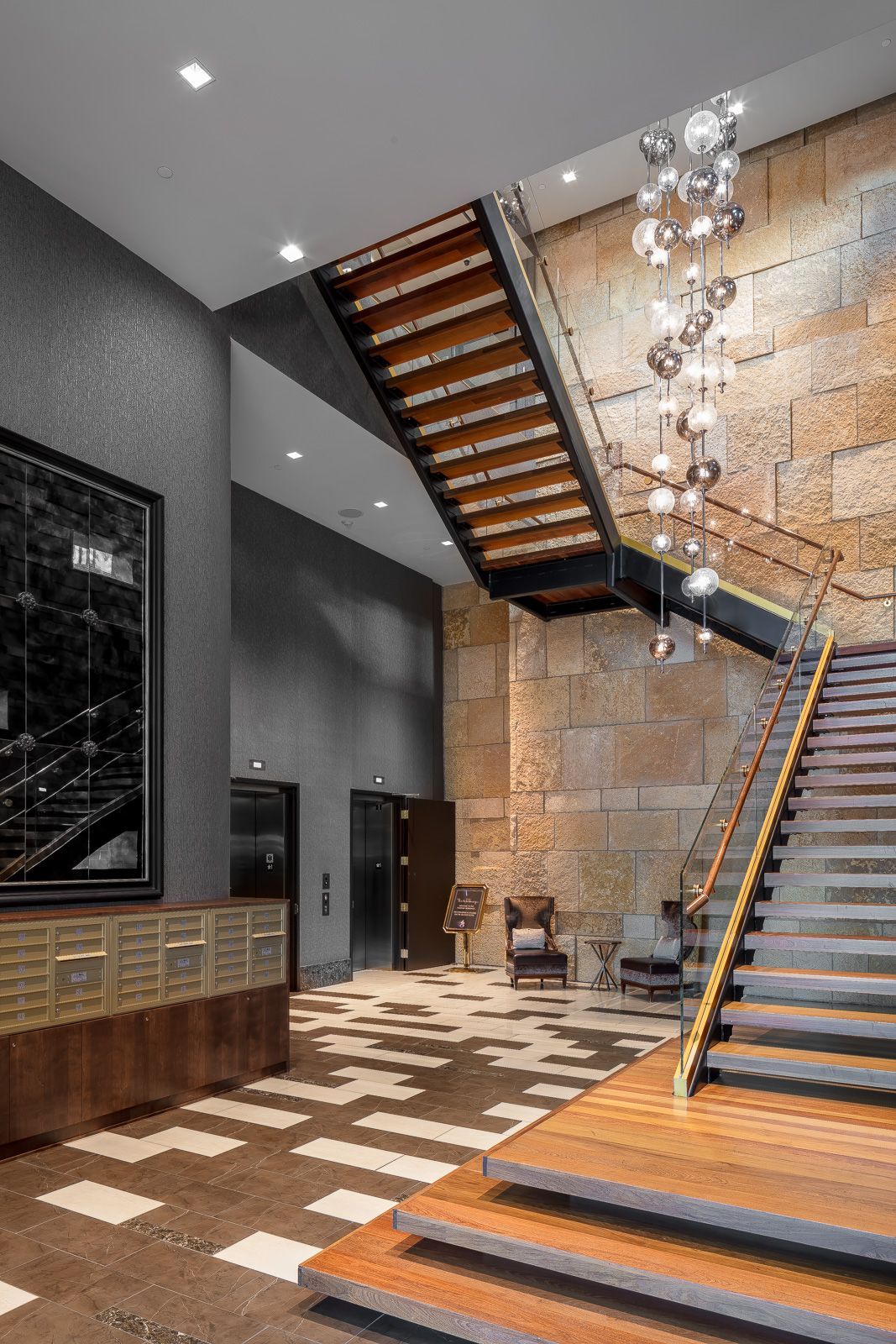 Modern apartment lobby with curved wooden staircase, stone wall, chandelier, and elevators at The Walkway, which offers apartments near downtown Minneapolis.