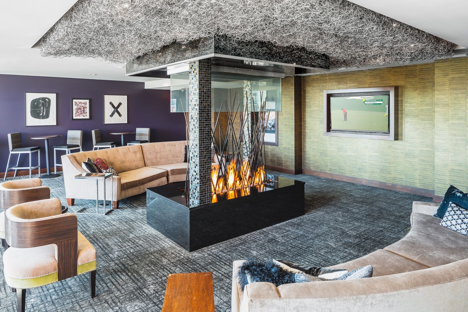 Lounge lobby with beige sectional seating, a central fire feature, purple accent wall, and a wall-mounted TV at The Walkway, which offers Minneapolis apartments for rent.