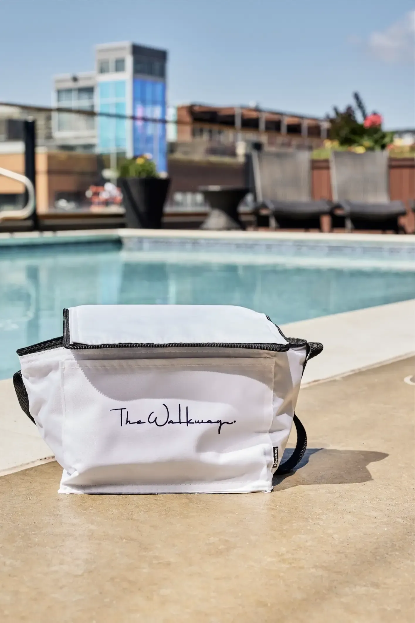 White bag with a black zipper sits on a pool deck; lounge chairs and a modern building are visible in the background at The Walkway, which offers apartments for rent in Minneapolis, MN.