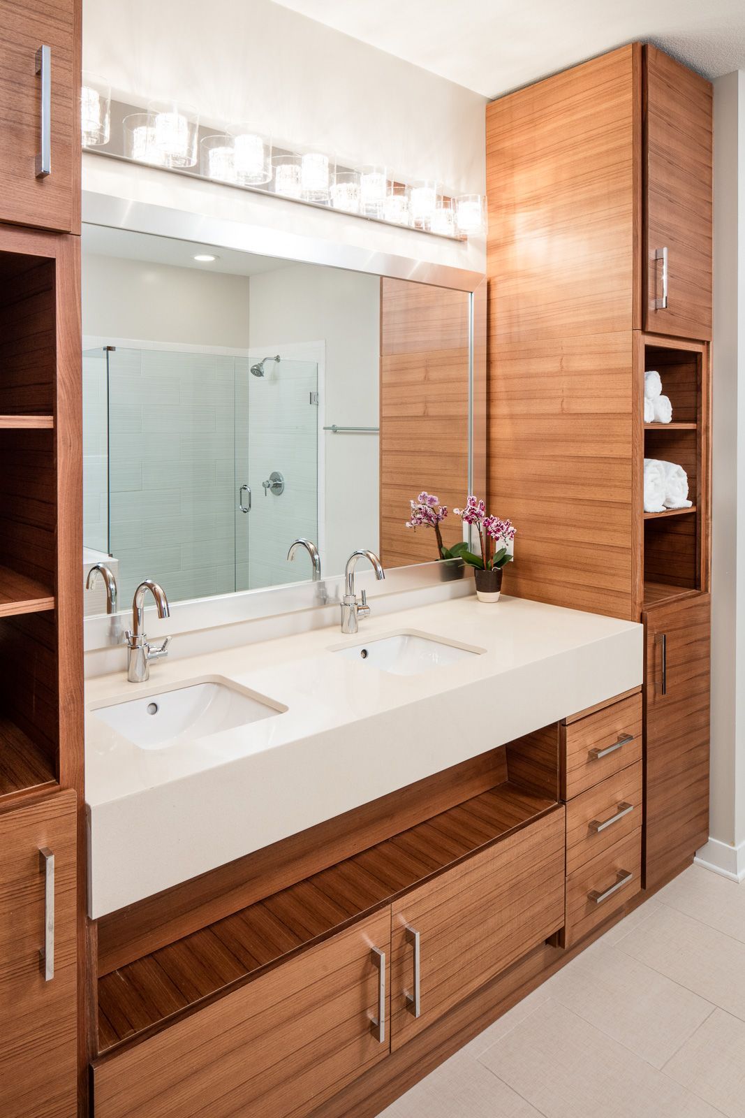Double-sink bathroom vanity with white countertop, wood cabinetry, and a glass-enclosed shower in the background at The Walkway, which offers Uptown apartments in Minneapolis, MN.