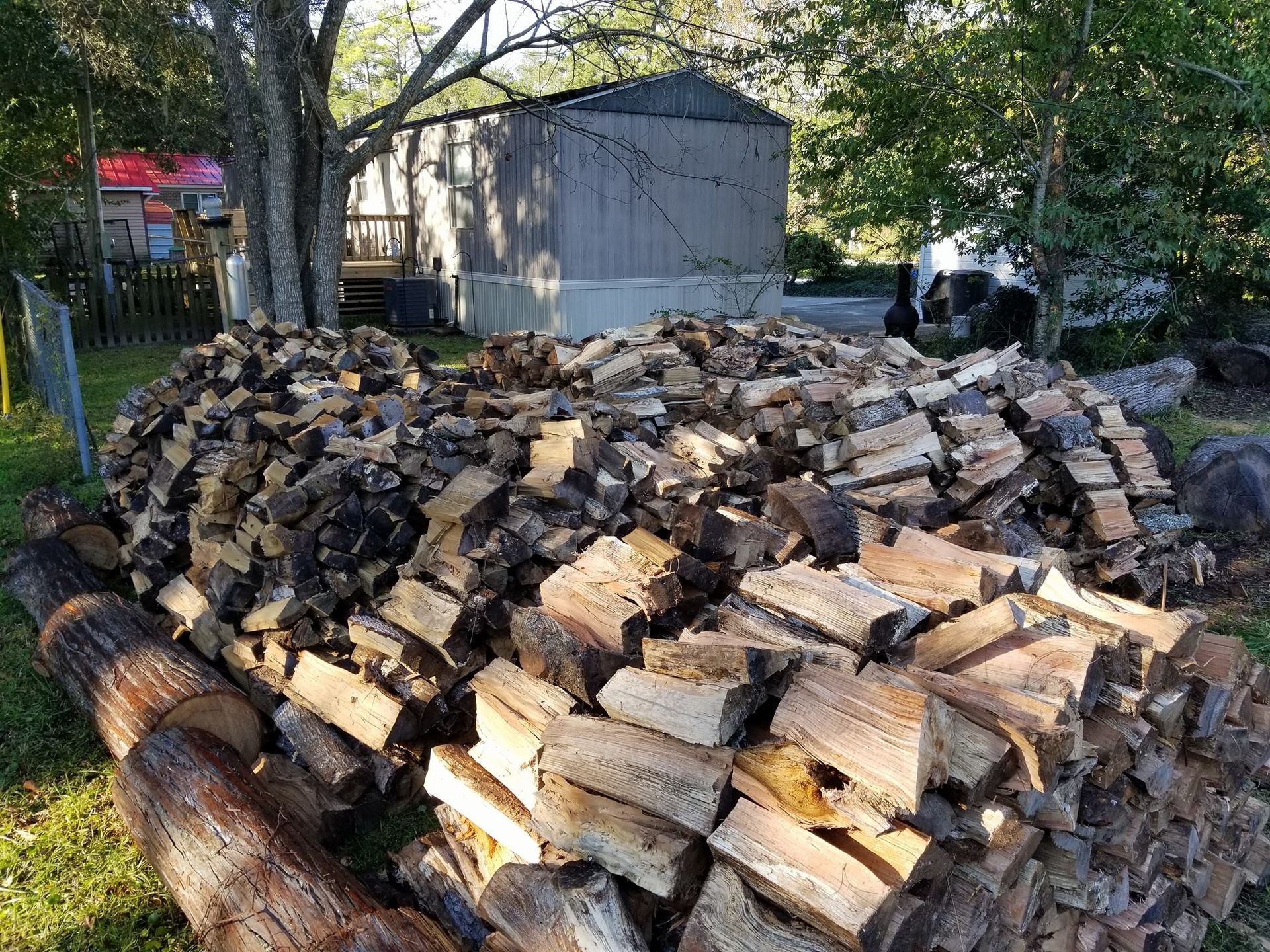 A large pile of logs is sitting in the grass in front of a shed.