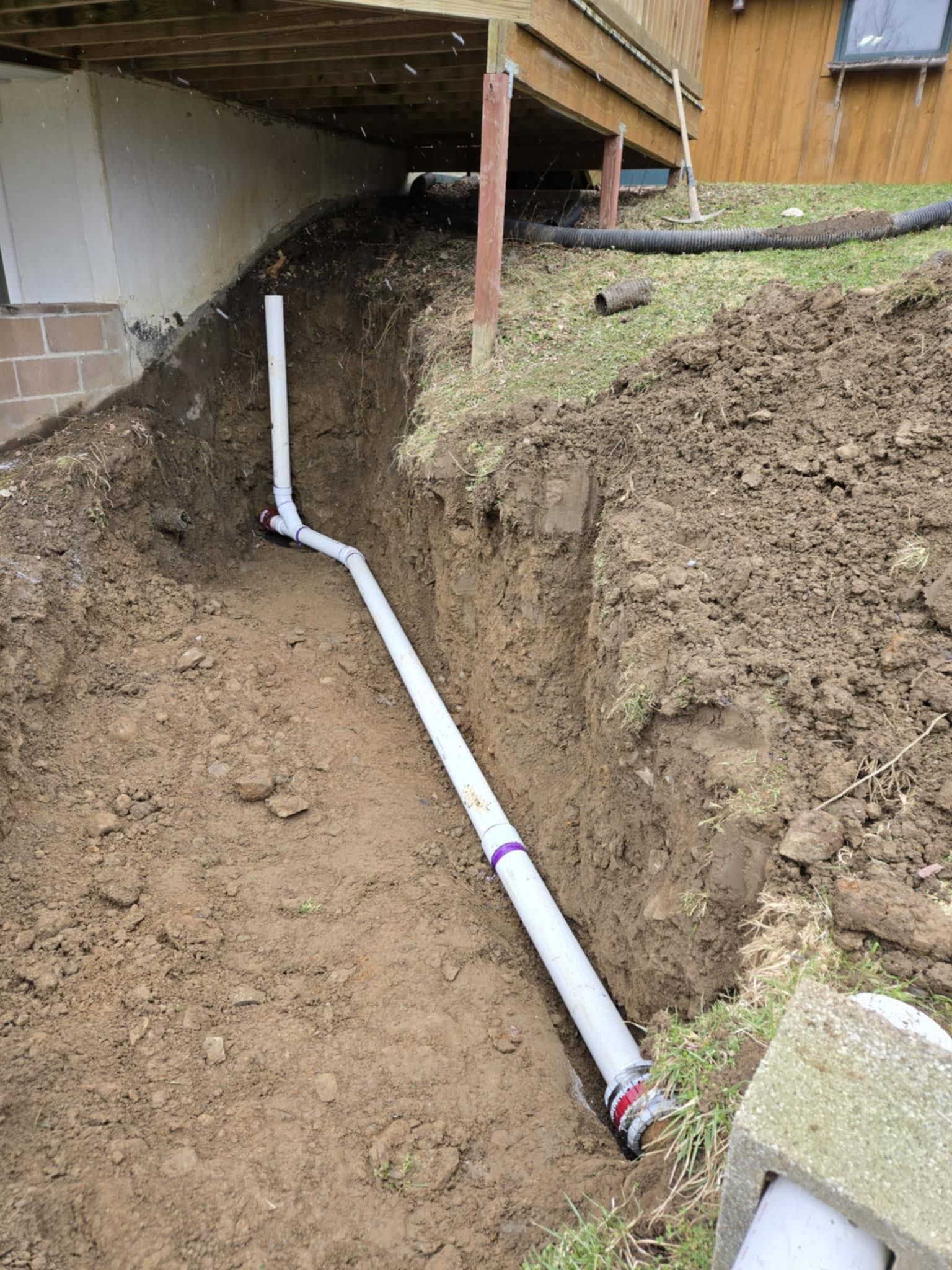 PVC drain pipe installed in a dirt trench beside a deck foundation.