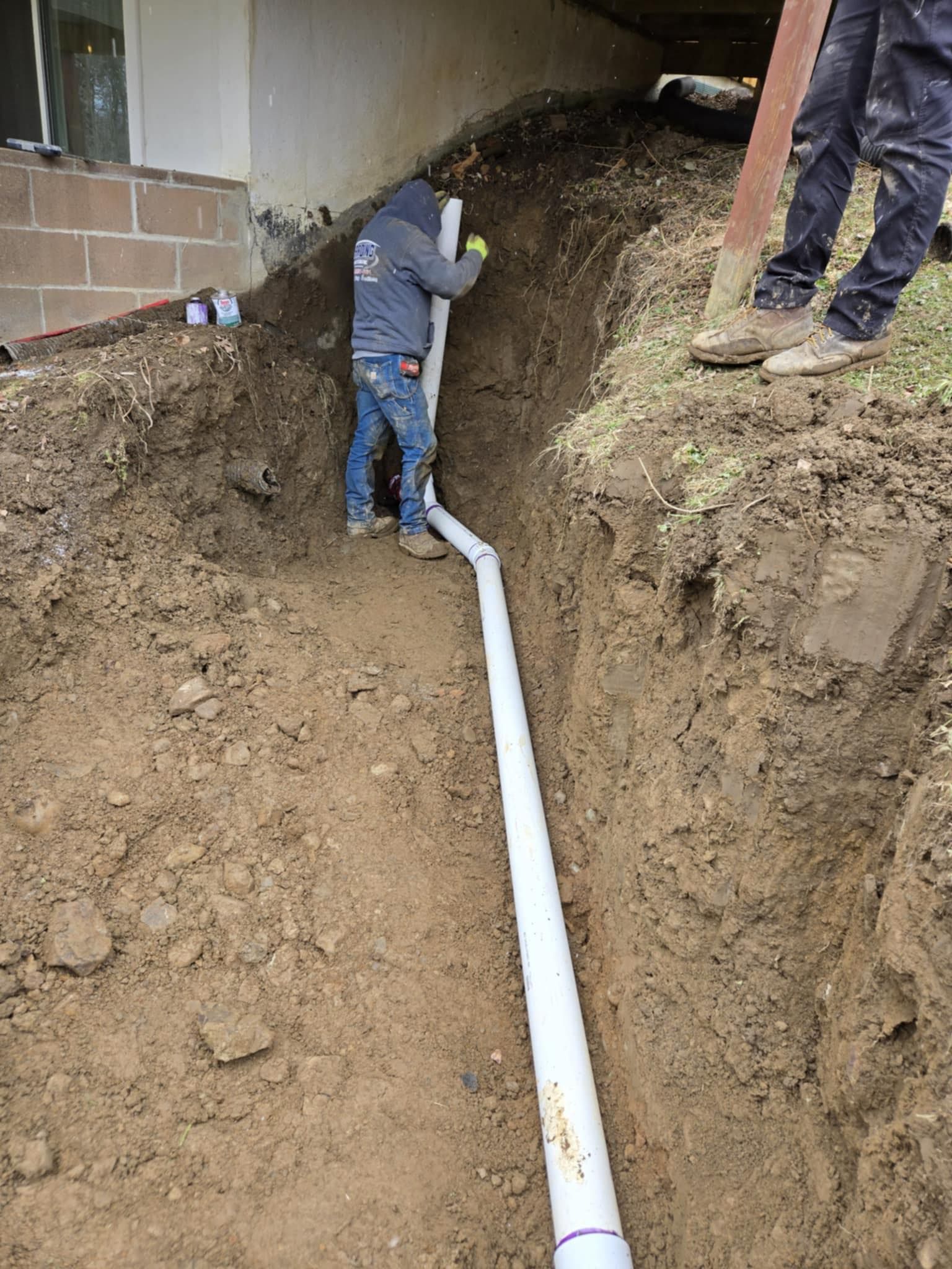 Workers installing a white pipe in a deep dirt trench beside a building