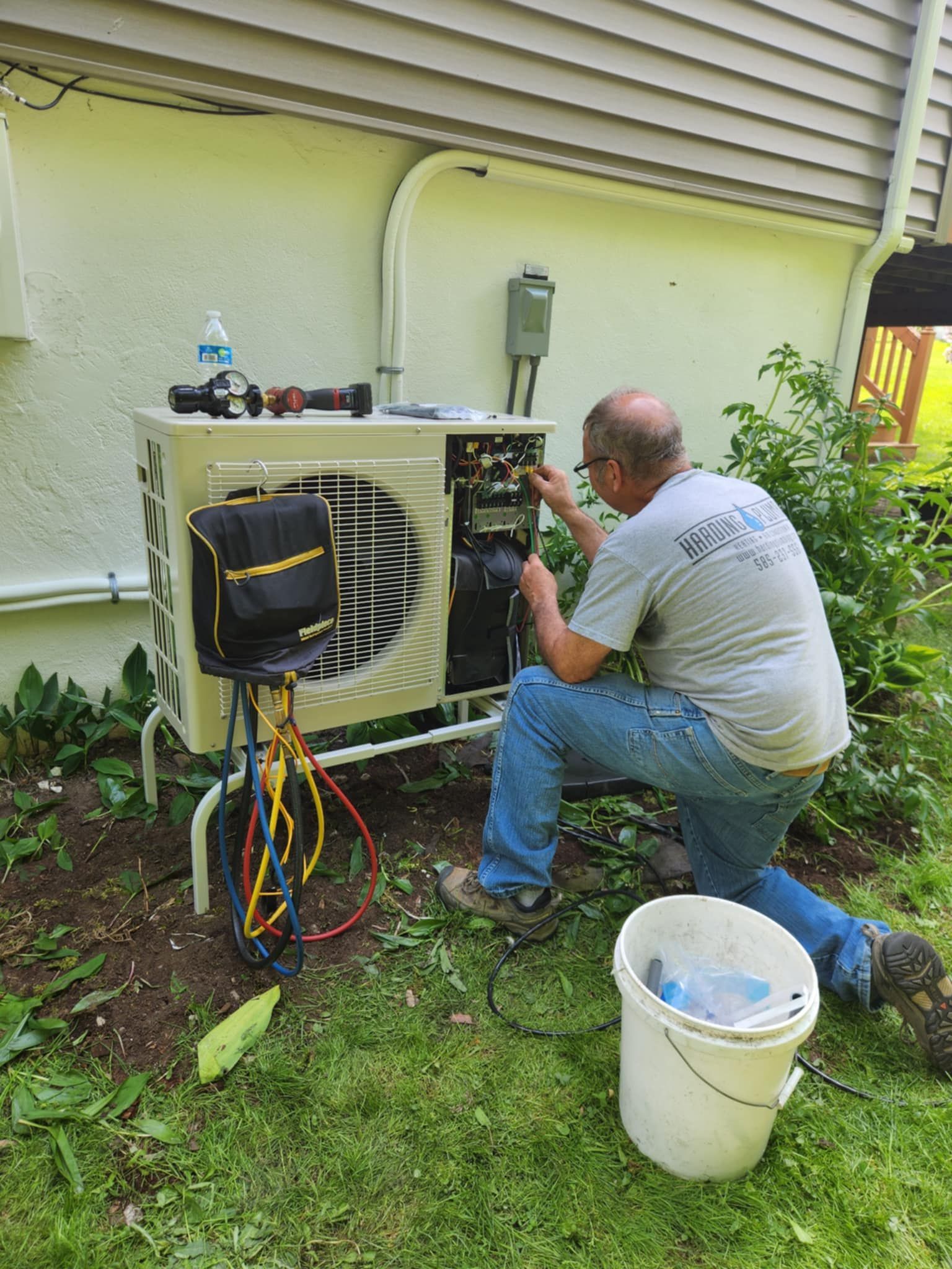 Technician servicing an outdoor HVAC unit beside a house, with tools and hoses on the ground.