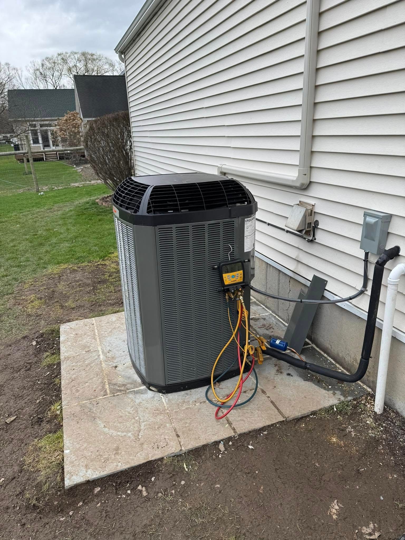 Outdoor HVAC unit beside a house with a hose and electrical cables attached, on a concrete pad.