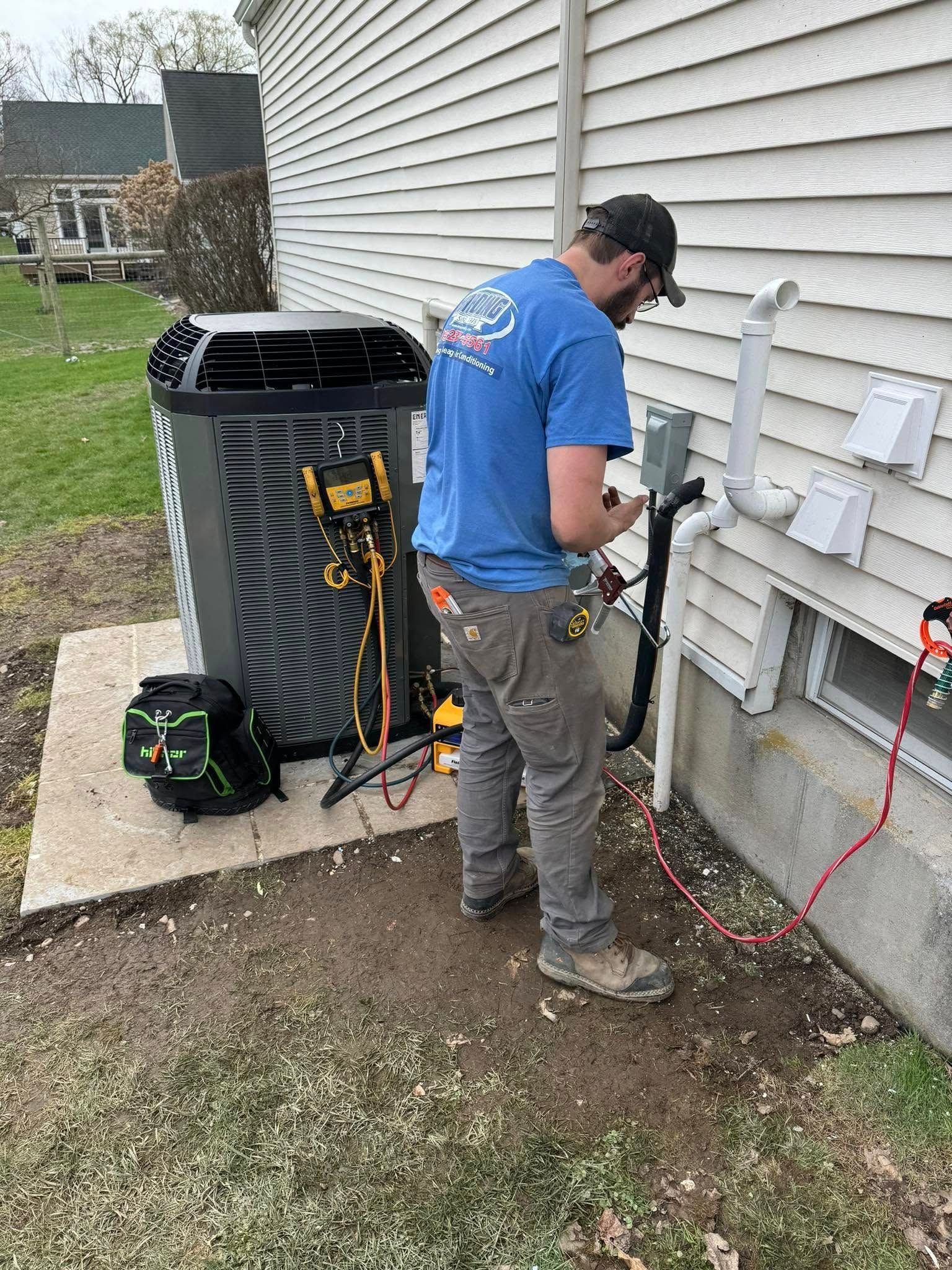 Technician servicing an outdoor HVAC unit beside a house, with tools and electrical connections visible.