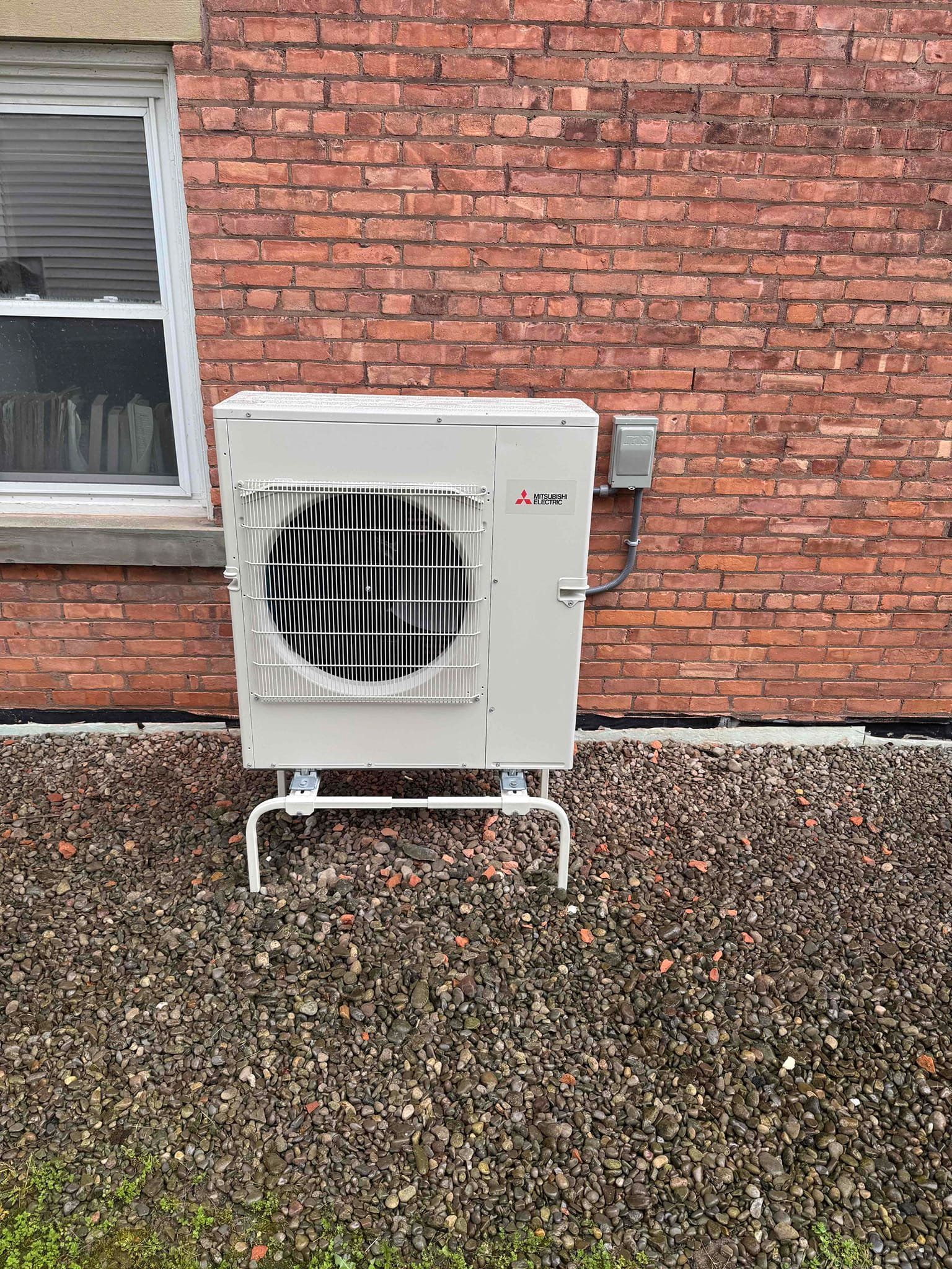 Outdoor air conditioning unit mounted on a brick wall beside a window, above gravel ground.