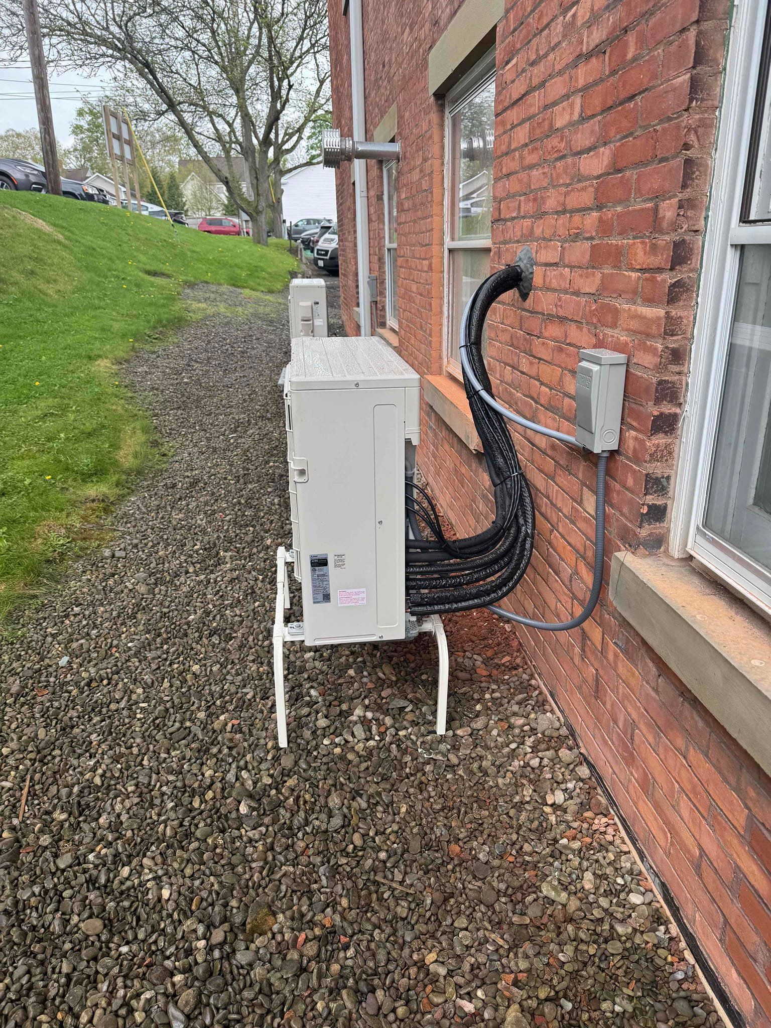 Outdoor HVAC unit beside a red brick house with black insulated piping and gravel ground