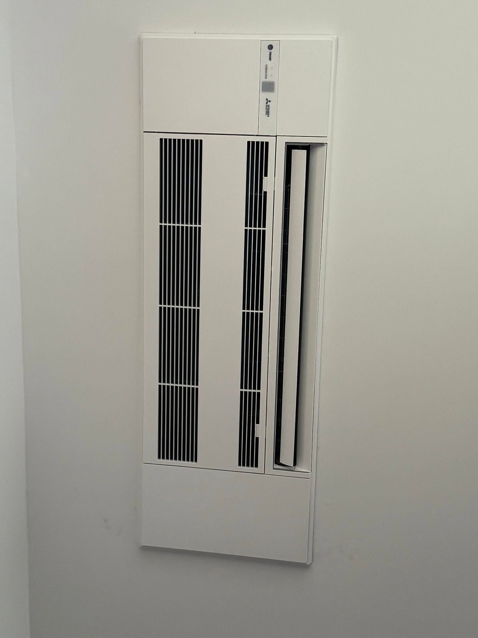 White wall vent cover with vertical black slats and bent center panel on a light gray wall