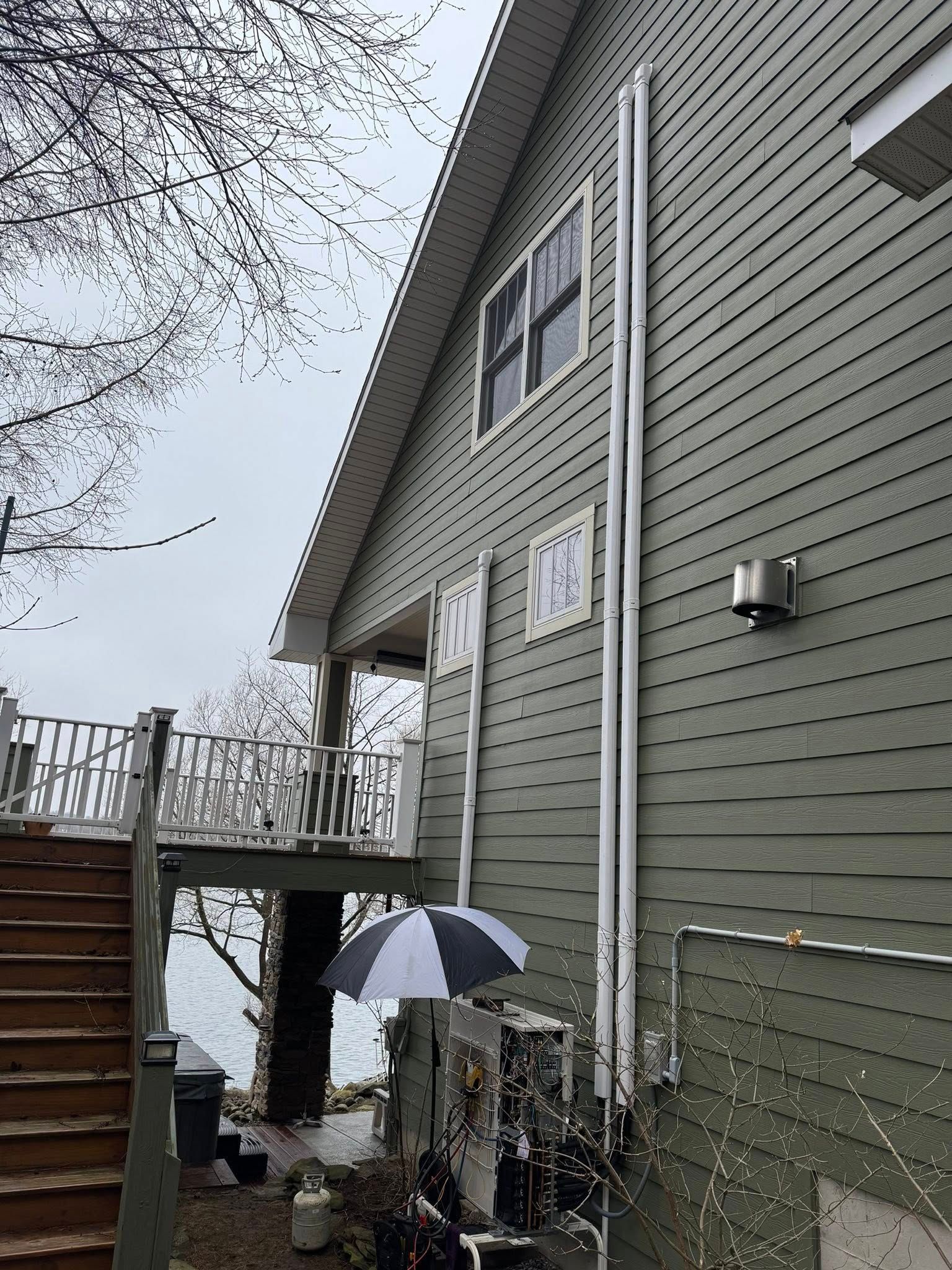 Snowy side view of a gray house with stairs, deck railing, and a striped umbrella by the lower entrance