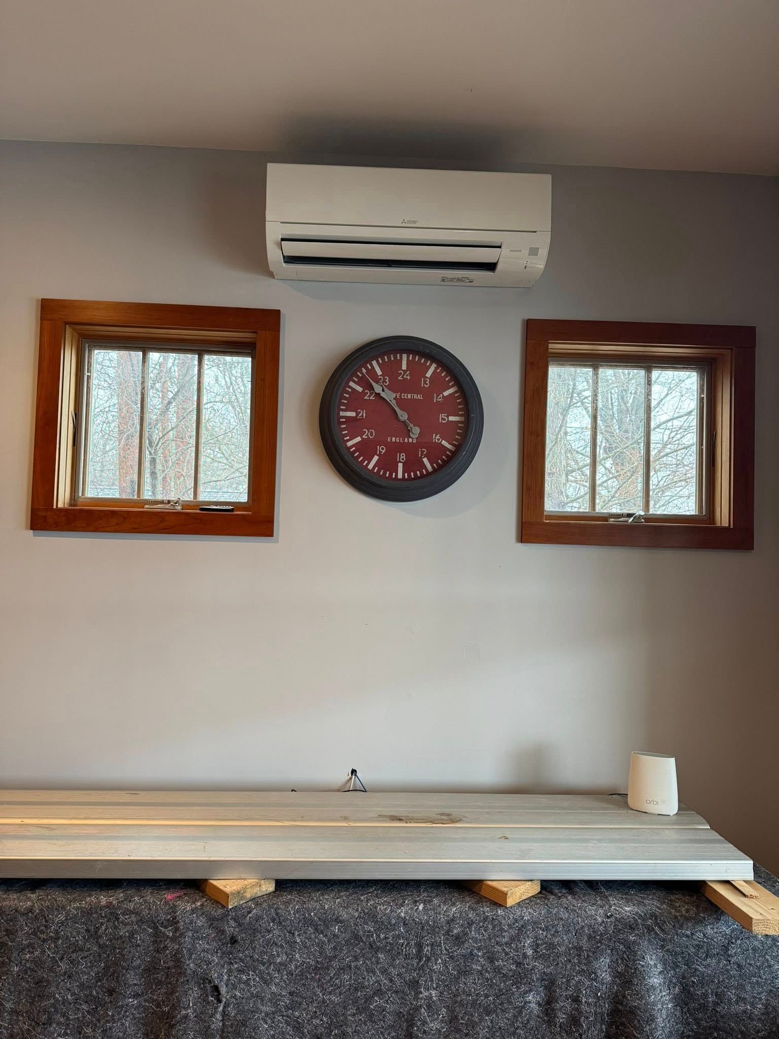 White wall with air conditioner, red clock, two wooden windows, and a counter or bench below.