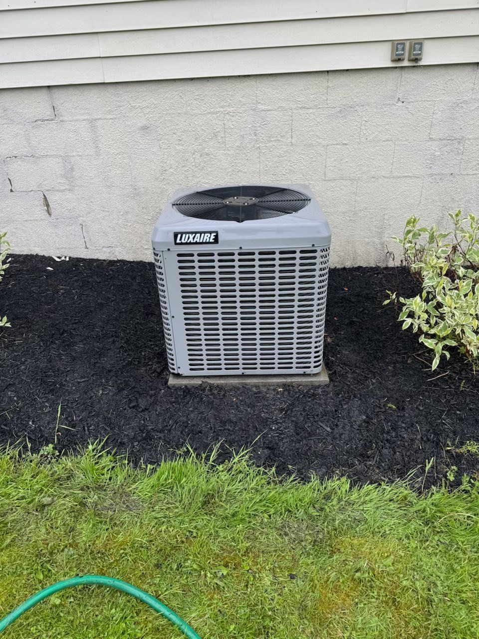 Outdoor air conditioner unit beside a house on a mulch bed with grass in front