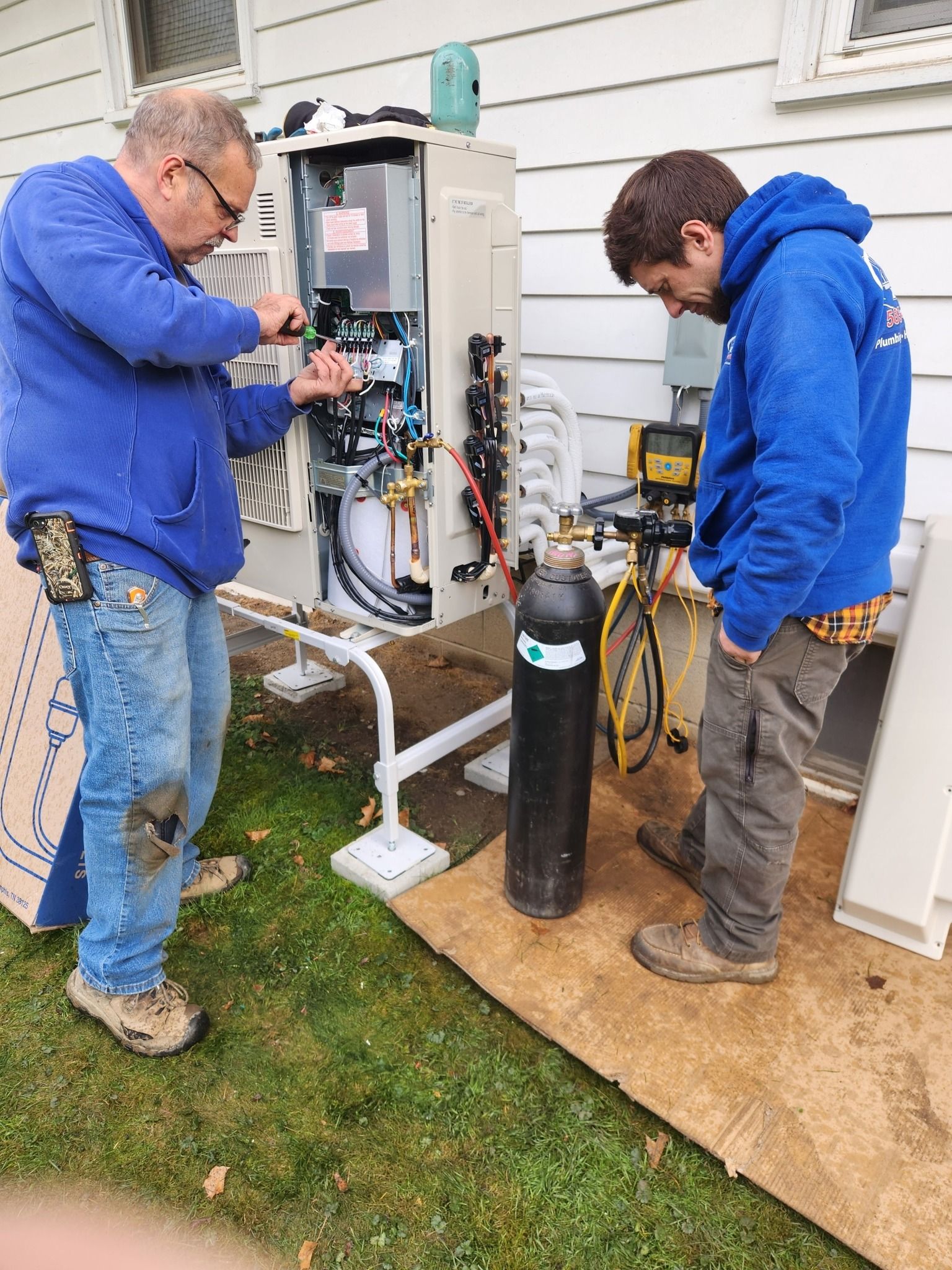 Two technicians work on an outdoor HVAC unit beside a house, with tools and gas cylinders nearby.