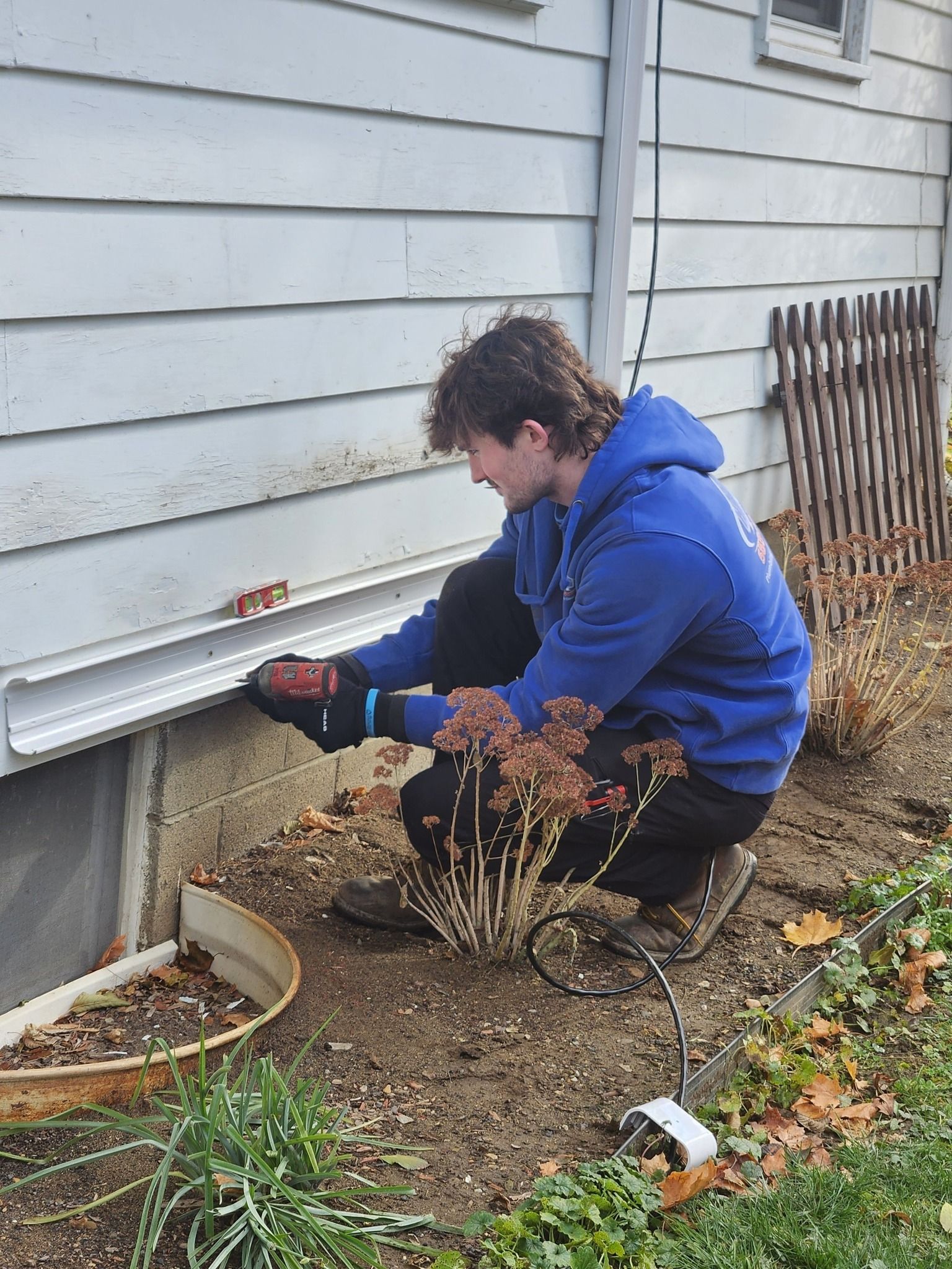 Person in blue hoodie cleaning debris from a house wall with a power tool in a yard