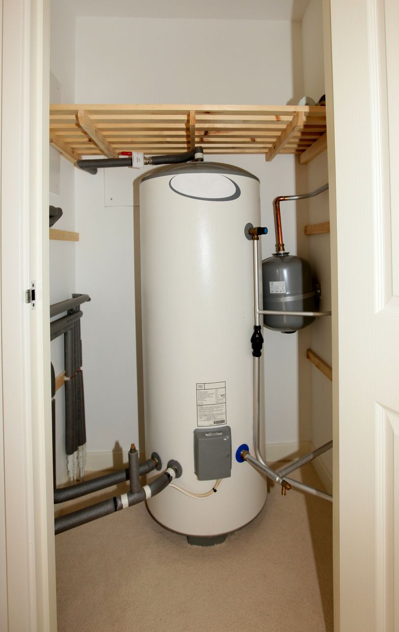 Water heater in a small closet with shelves, pipes, and a wall-mounted tank.