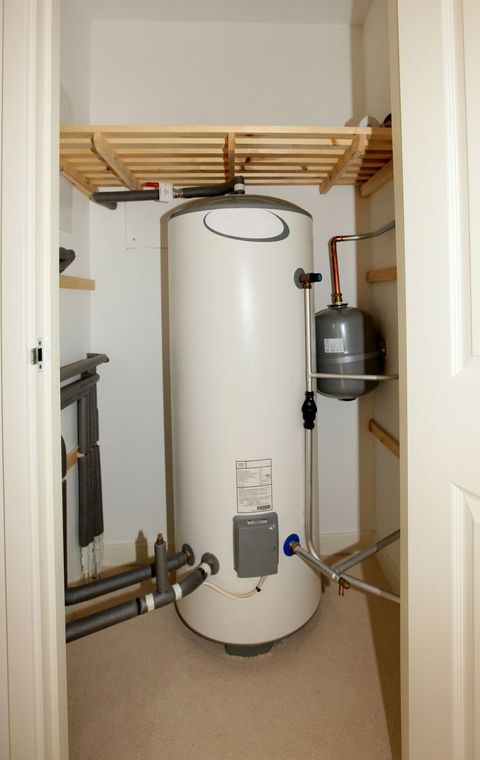 Water heater in a small closet with shelves, pipes, and a wall-mounted tank.