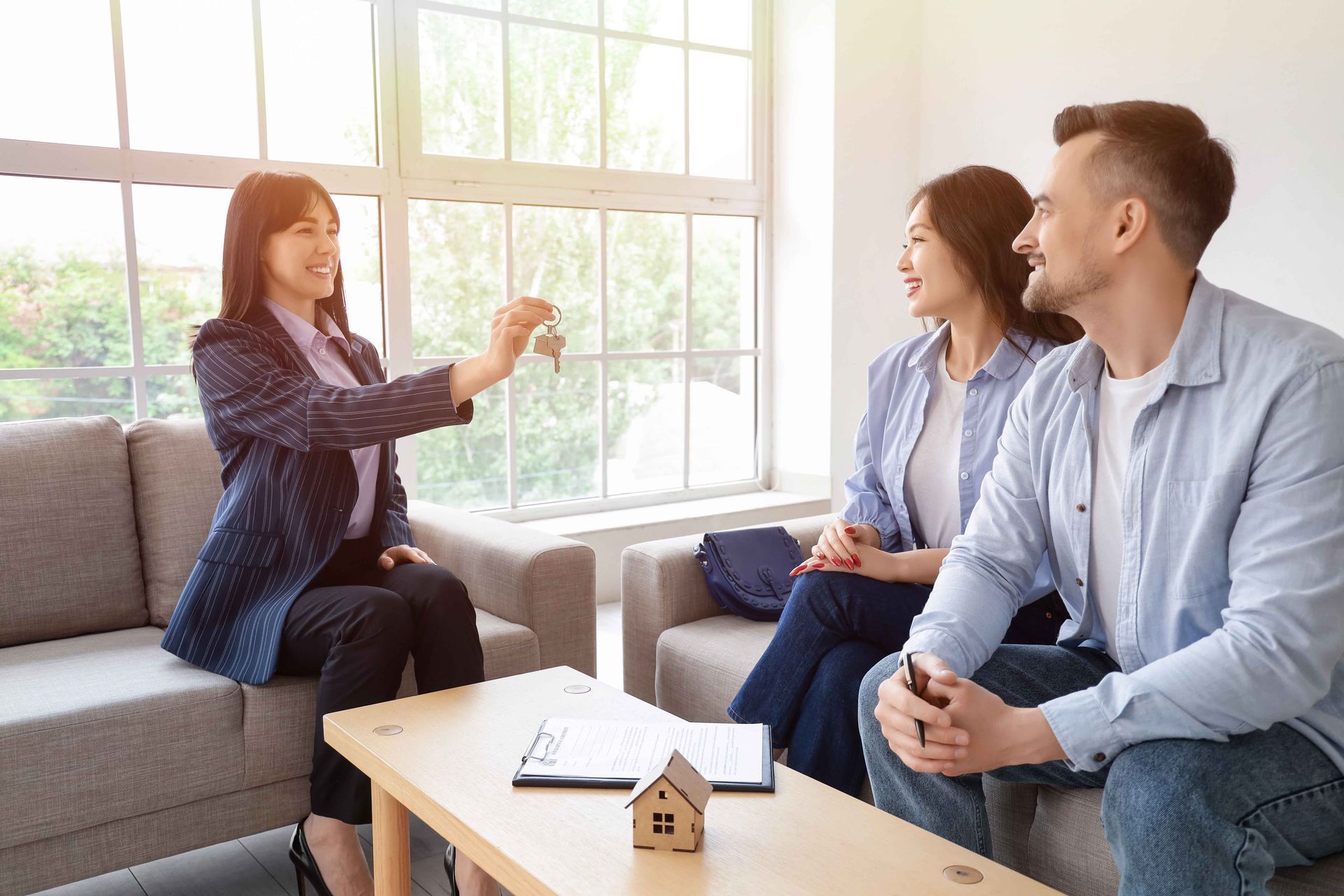Un agent immobilier remet les clés à un couple dans un salon lumineux, qui signe des documents sur une table.